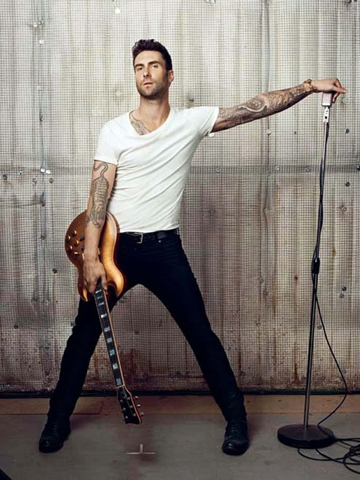 Maroon 5主唱亞當·萊文 壁紙 — 一位紋身男子身穿白色T恤和黑色長褲，在生鏽的金屬牆前自信地擺姿勢，手持結他和咪高峰。