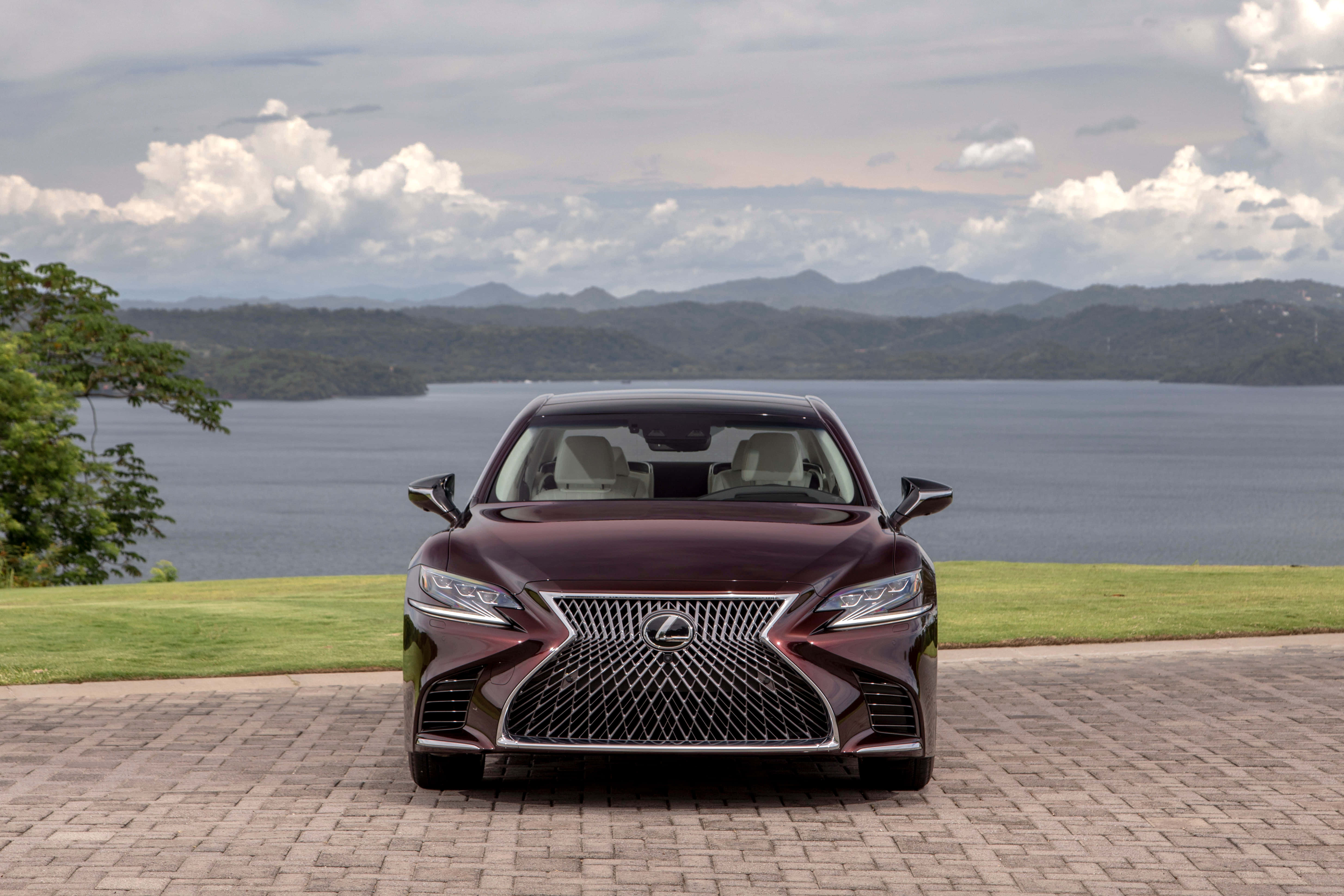 Lexus Ls 500 桌布 壁紙 — 一輛栗色Lexus房車在磚砌車道上向前行駛，前方是廣闊的湖泊和森林覆蓋的山脈，天空多雲。