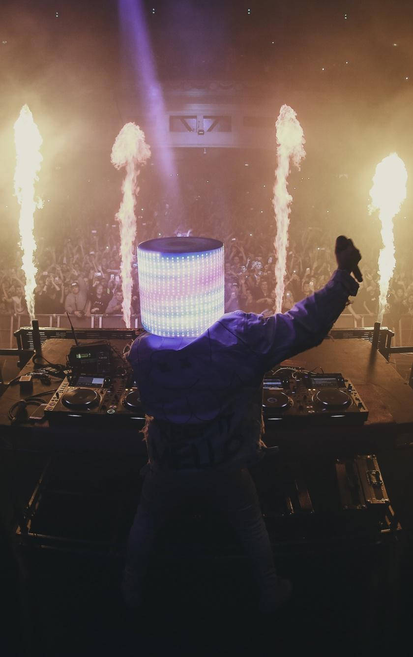 Marshmello iPhone 演唱會歡樂 壁紙 — 一位戴著發光LED頭飾的DJ在舞台上充滿活力地表演，身後有熾熱的煙火爆炸。