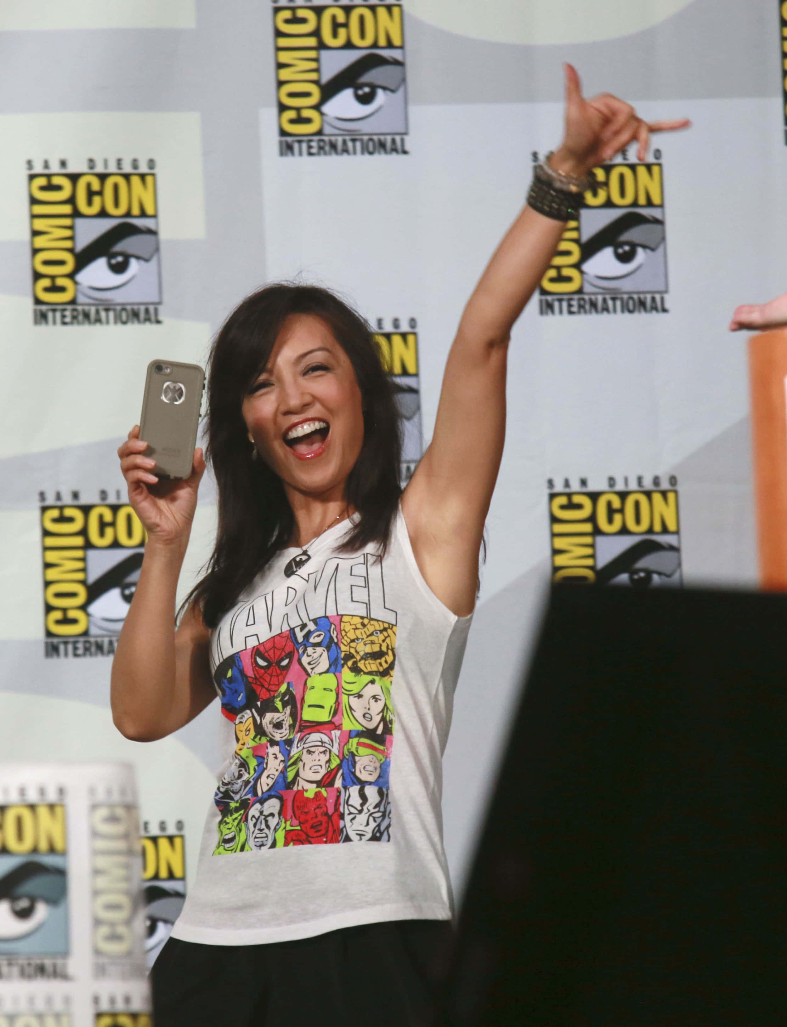 Ming Na Wen 桌布 壁紙 — 一位身穿Marvel T恤的快樂女性，在Comic-Con活動中舉起手機並做出和平手勢，露出燦爛的笑容。