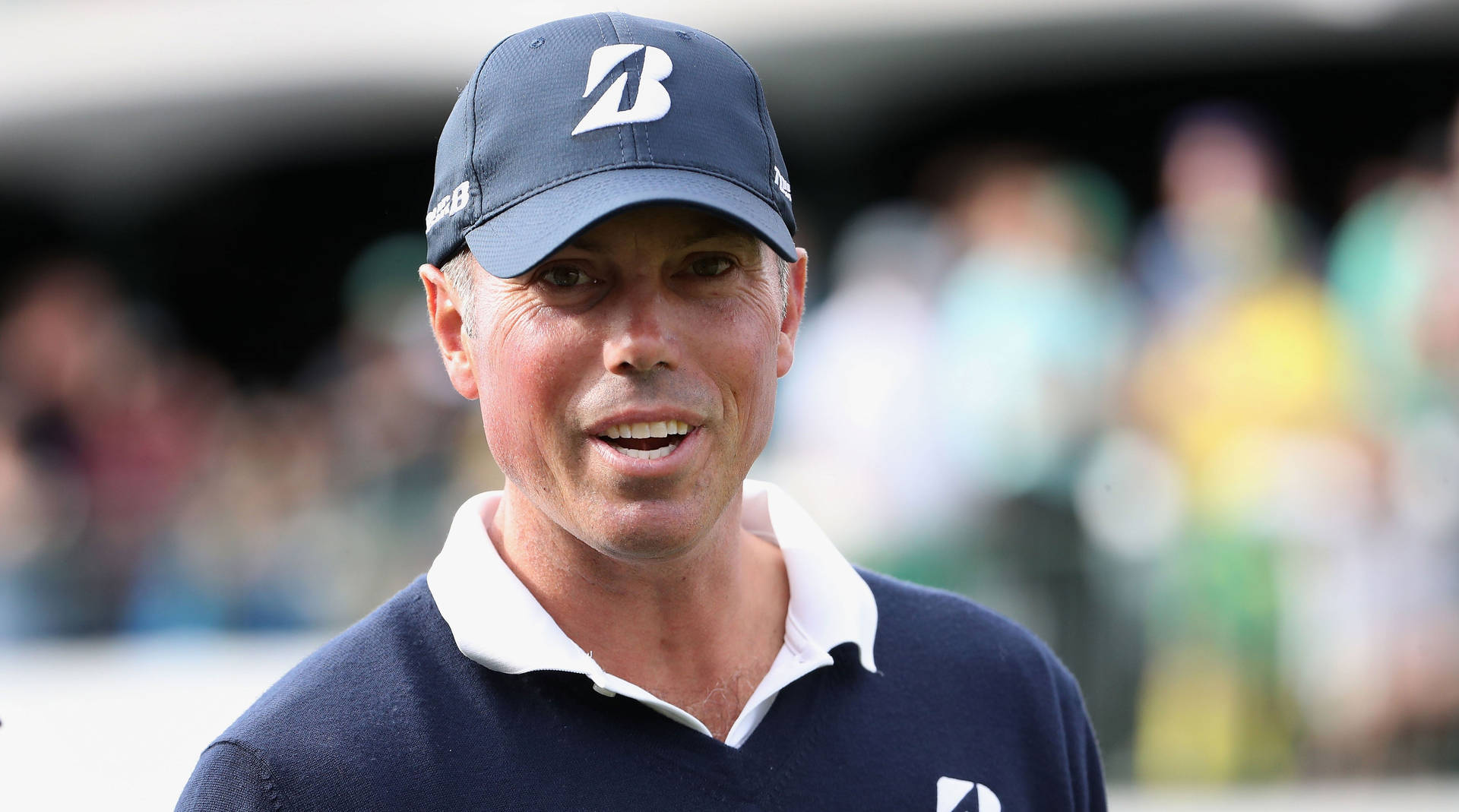 Matt Kuchar 微笑特寫 壁紙 — 一位專注的高爾夫球手戴著海軍藍帽子和Polo衫，在日間比賽中微笑，身後有模糊的觀眾。