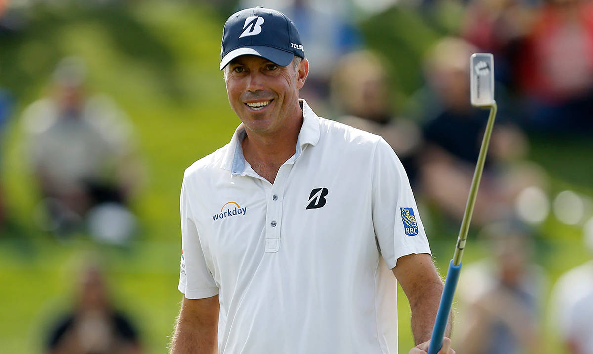 Matt Kuchar 握著高爾夫球桿 壁紙 — 一位微笑的高爾夫球手，身穿白色Polo衫和帽子，在陽光明媚的高爾夫球場上握著球桿，背景有模糊的觀眾。