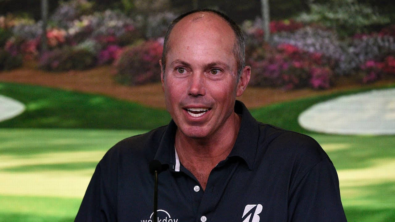 Matt Kuchar 頭髮稀疏特寫 壁紙 — 一位微笑的光頭男子穿著深色Polo衫，在充滿活力的高爾夫球場背景下於新聞發布會上發言。