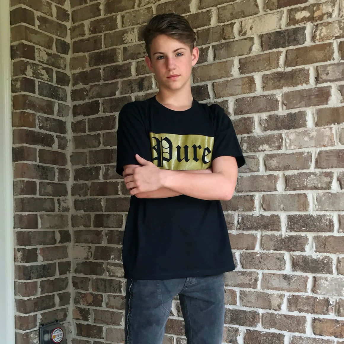 身穿黑色T恤和牛仔褲的青少年男孩，交叉雙臂靠著磚牆站立。