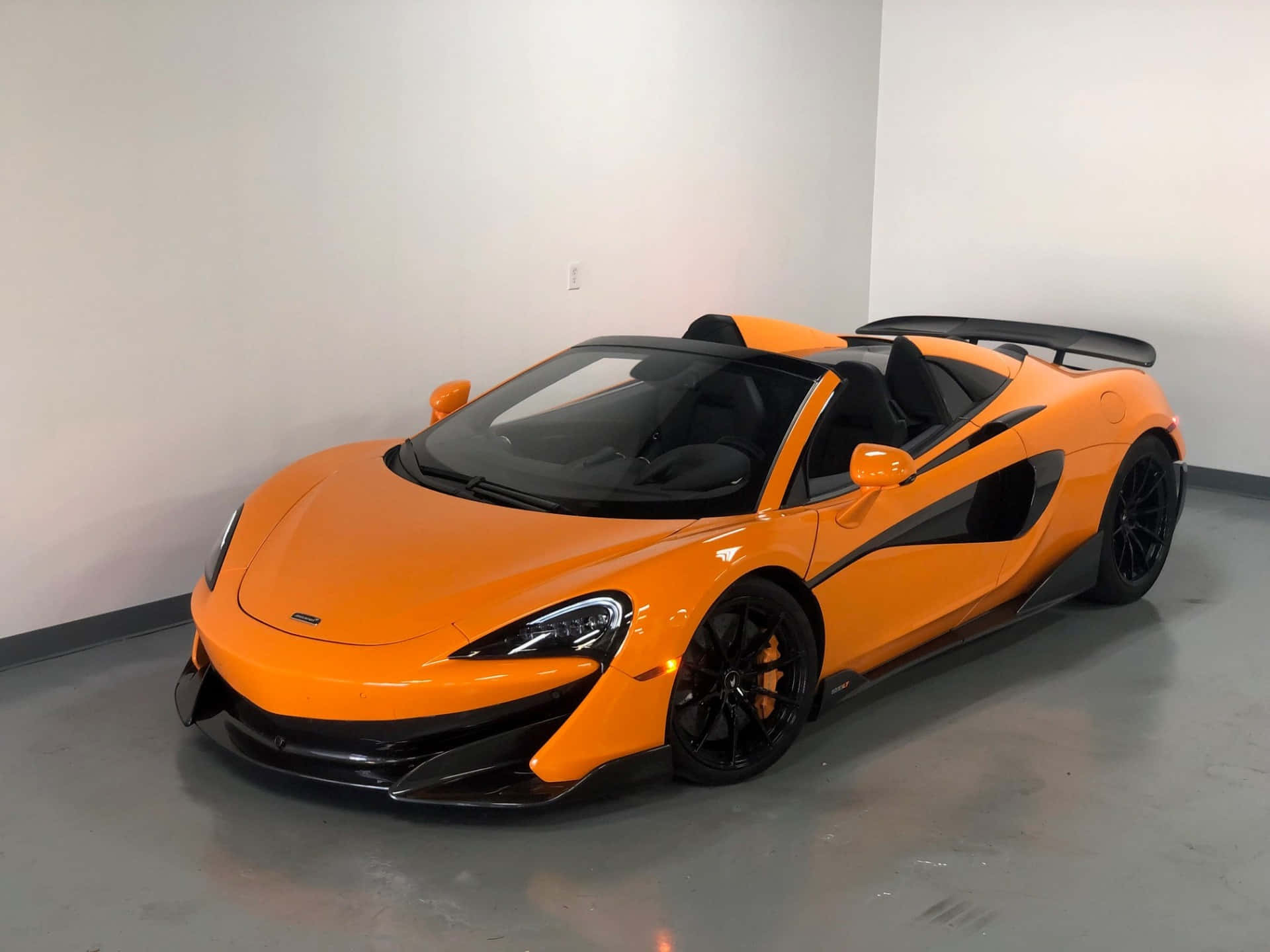 Mclaren 600lt Spider 桌布 壁紙 — 一輛充滿活力的橙色McLaren 570S Spider，帶有黑色飾邊和可拆卸車頂，停在室內的灰色地板上。