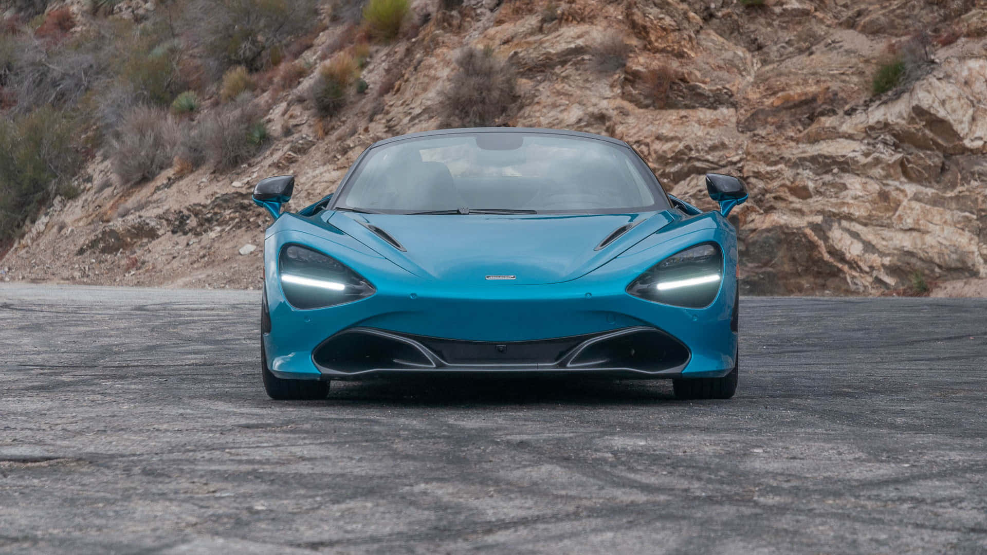 霓虹青色 McLaren 720S 壁紙 — 一架引人注目的青綠色McLaren 720S跑車在崎嶇的山路上向前行駛，背景是岩石峭壁和乾燥的灌木叢。