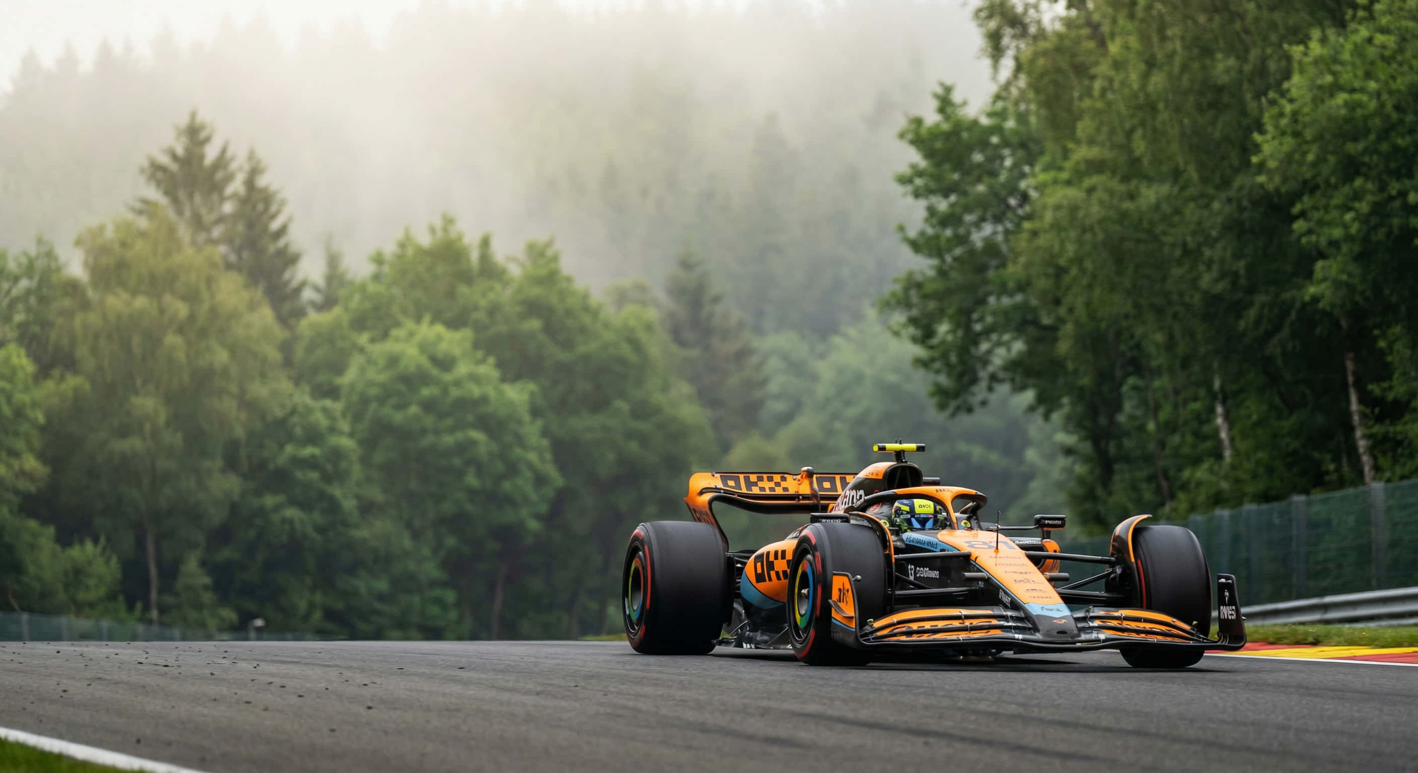 McLaren MCL-39（Oscar Piastri）賽車壁紙 壁紙 — 一輛鮮豔的橙色和黑色一級方程式賽車在潮濕的瀝青賽道上疾馳，賽道兩旁是茂密的綠色森林，天空霧氣瀰漫。