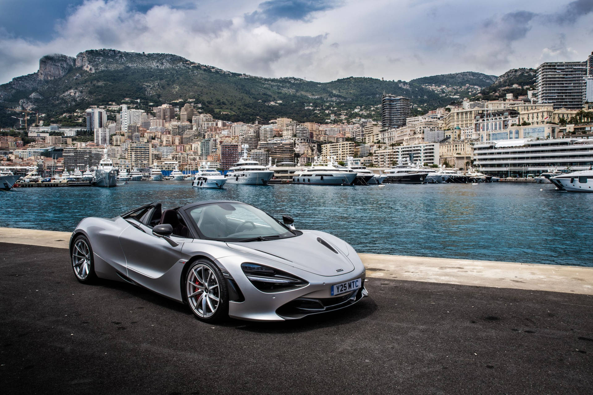 銀色McLaren 720S Spider停泊在遊艇碼頭，背景有遊艇和城市建築。