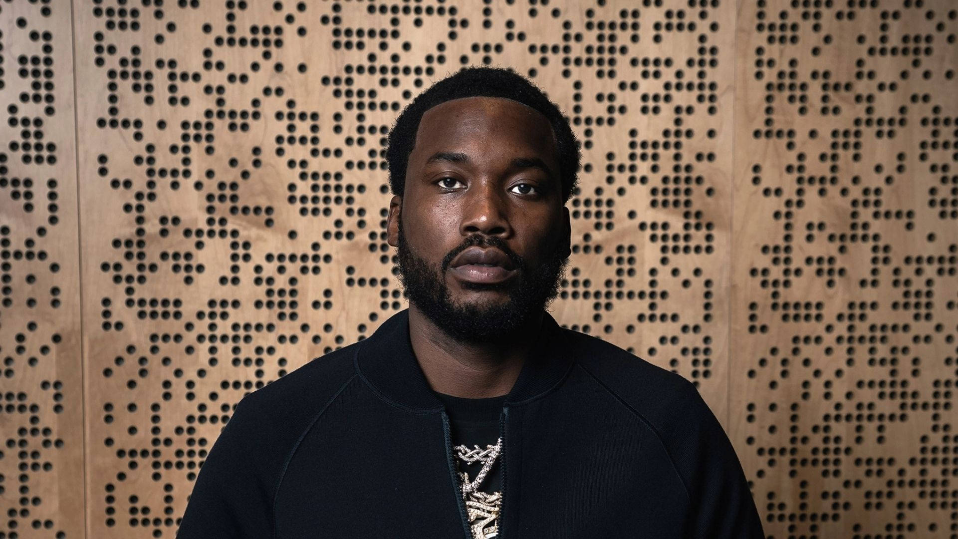 Meek Mill 試圖掩飾表情 壁紙 — 一個留鬍鬚的男人直視鏡頭，背景是帶有圓點幾何圖案的木牆。