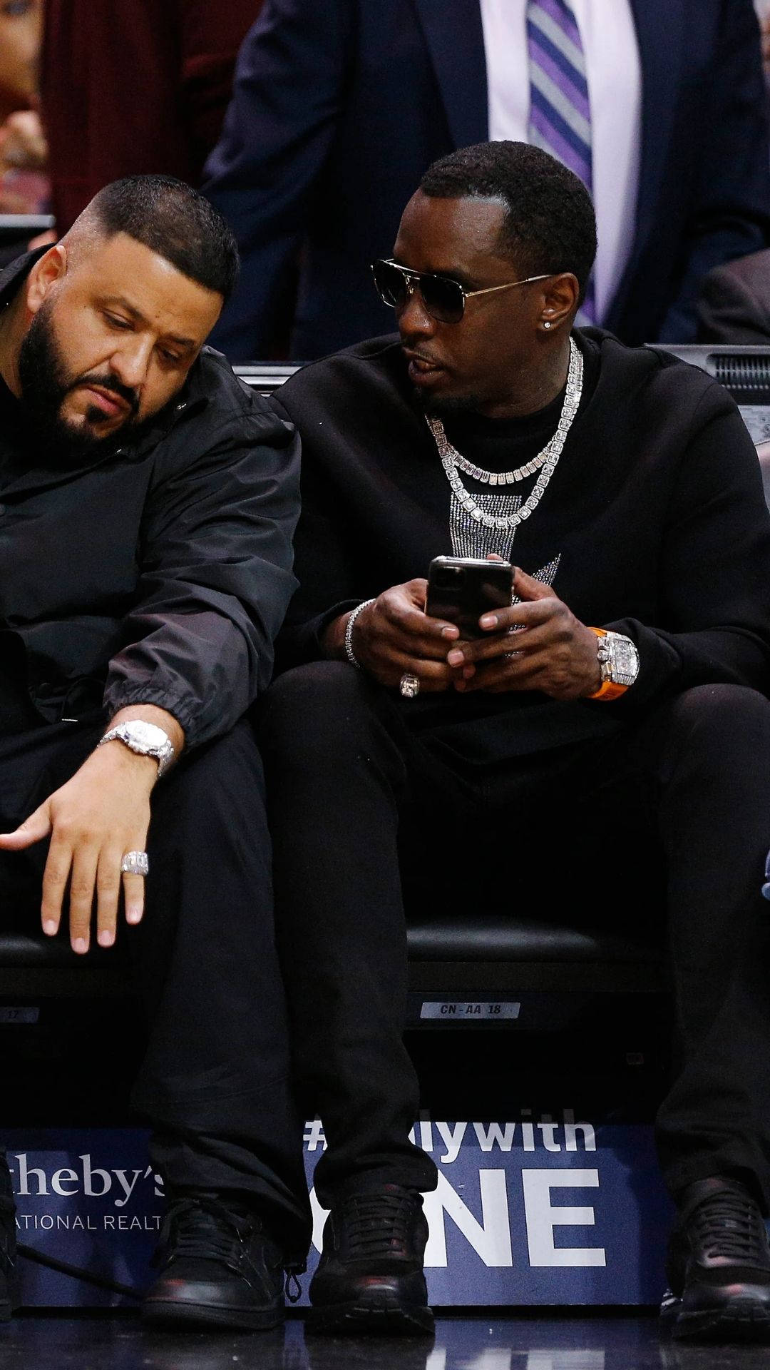 Meek Mill 與 DJ Khaled 壁紙 — 兩名身穿黑色服裝的男子坐在場邊，一人查看手機，另一人將手放在膝蓋上，兩人都佩戴著奢華珠寶。