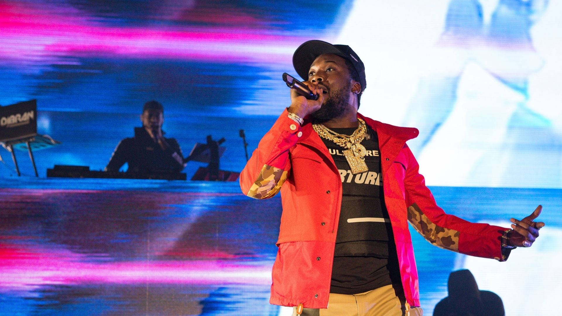 Meek Mill 唱歌 壁紙 — 一位身穿紅色外套的饒舌歌手在舞台上表演，背景有DJ，舞台燈光為鮮豔的粉紅色和藍色。