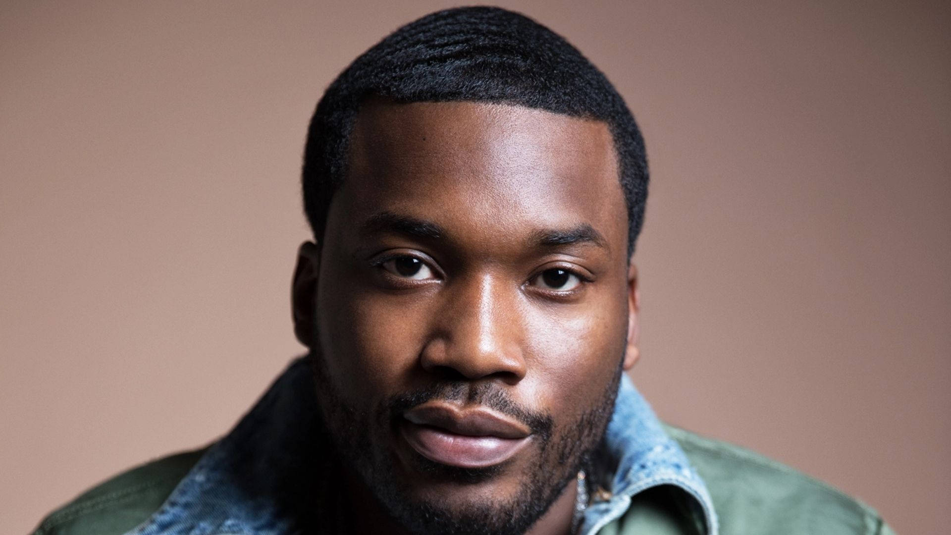 Meek Mill 嘲笑式微笑 壁紙 — 一位黑人的特寫肖像，短髮留鬍，身穿牛仔布領外套，背景為柔和的棕色。