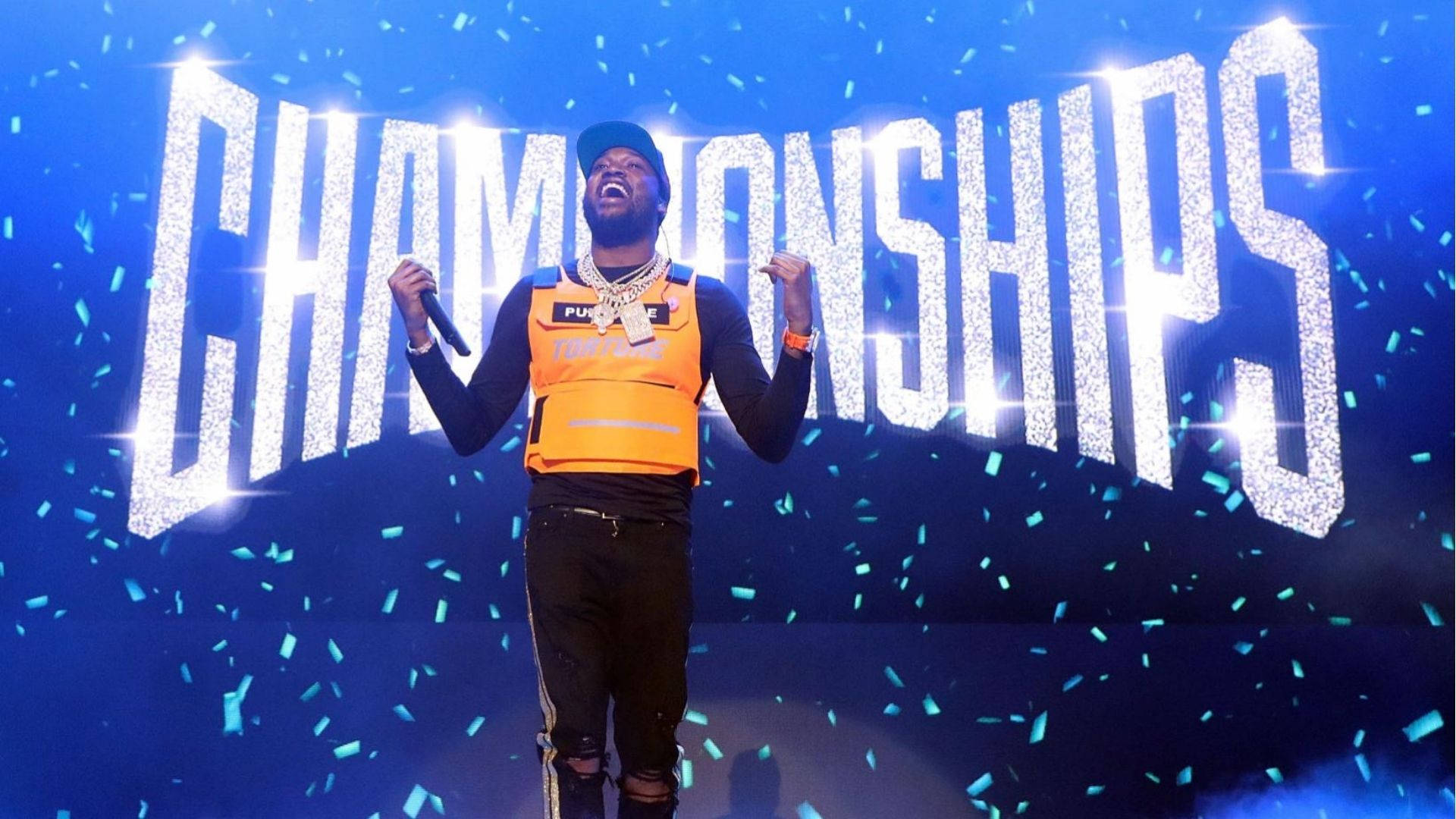 Meek Mill 在紐約 壁紙 — 一位穿著橙色背心和黑色帽子的說唱歌手在舞台上，在閃閃發光的「CHAMPIONSHIPS」標誌下，藍色紙屑飄落，慶祝勝利。