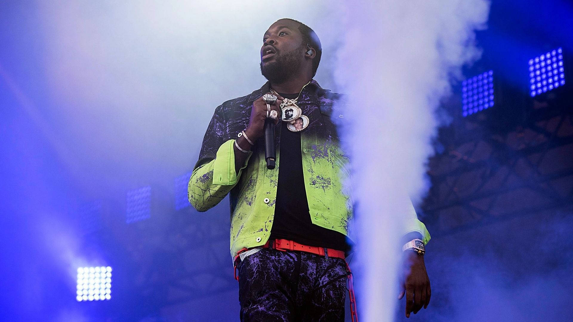 Meek Mill 在霧氣舞台 壁紙 — 一位身穿霓虹外套的男性表演者，在煙霧繚繞、藍色聚光燈照射的舞台上對著麥克風唱歌。