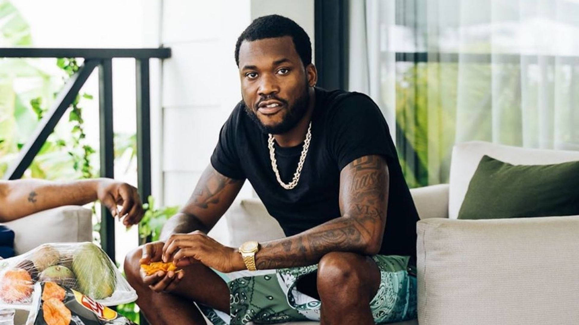 Meek Mill 度假 壁紙 — 一個有紋身同金鏈嘅男人隨意咁坐喺白色梳化上食生果，同時望住鏡頭。