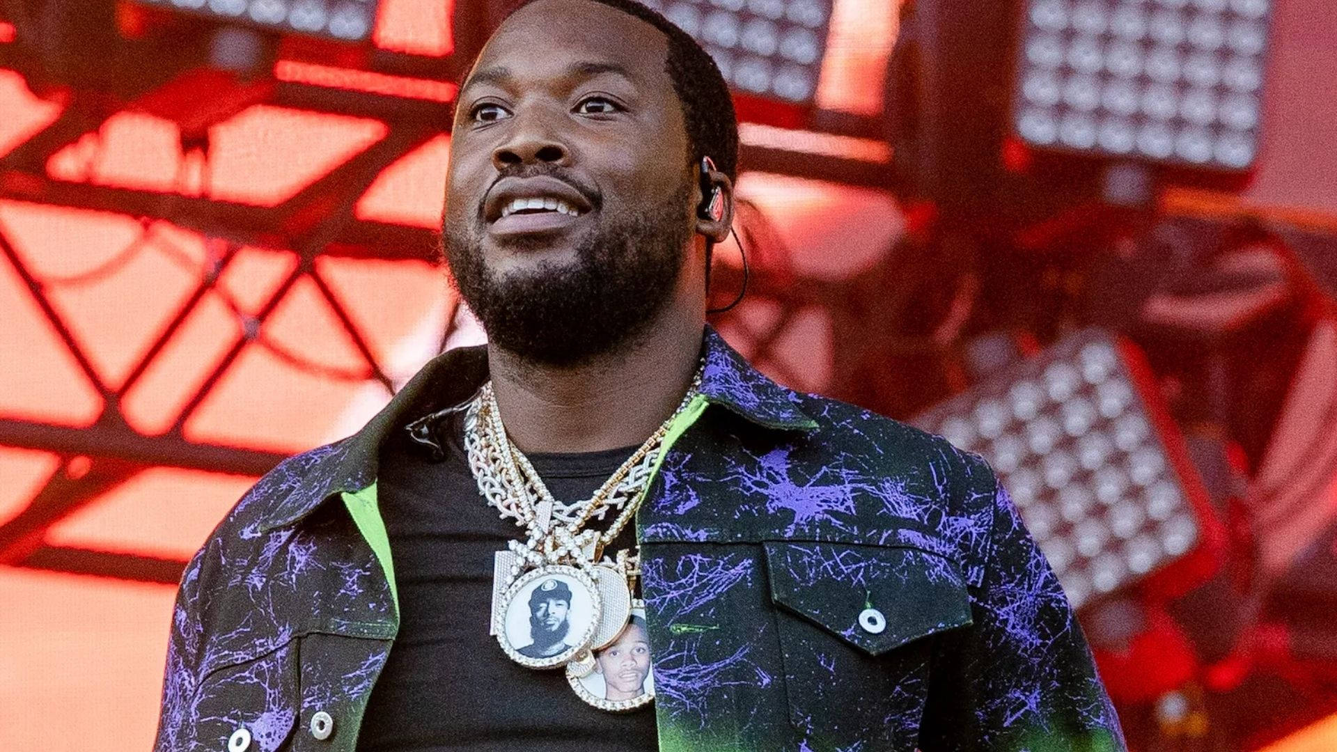 Meek Mill 微笑對著粉絲 壁紙 — 一位留鬍鬚的 smiling 男士穿著圖案外套和層疊金鏈，背景是紅色舞台和燈光。