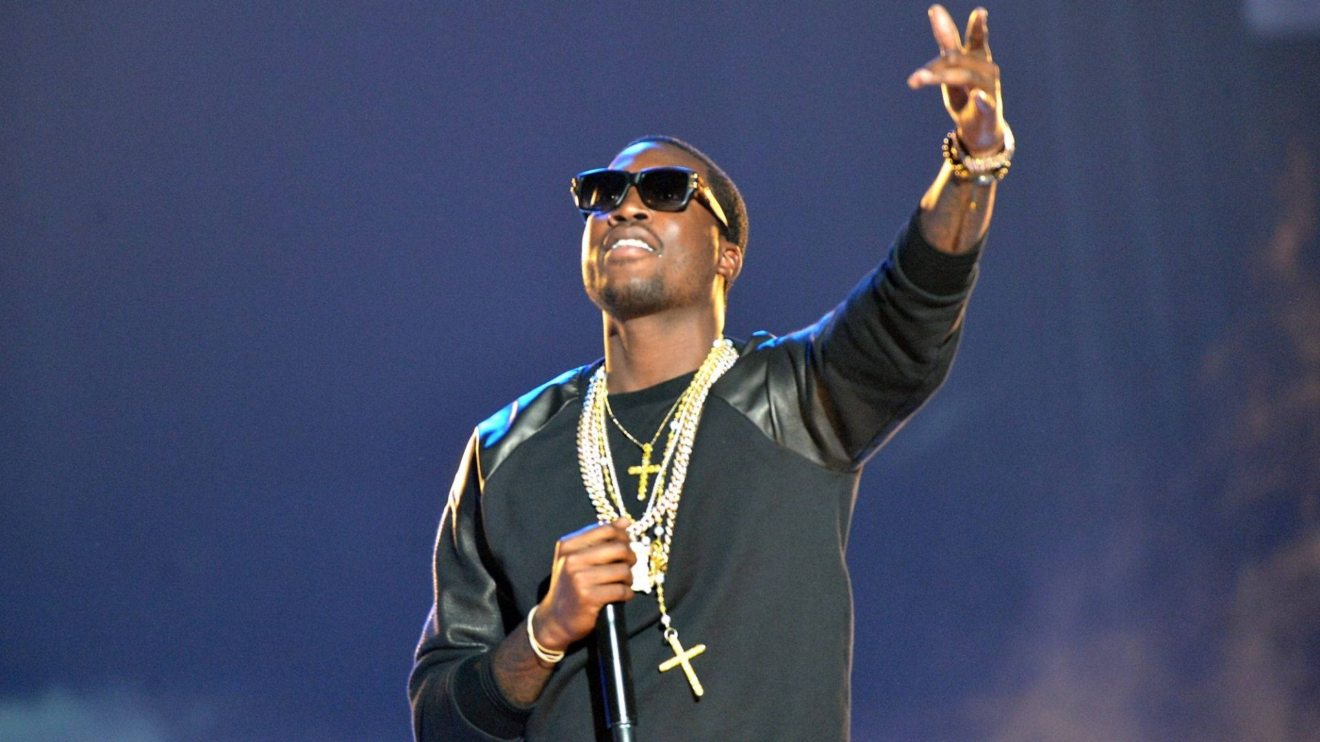 Meek Mill 戴太陽眼鏡 壁紙 — 一位戴著太陽眼鏡和金鏈的饒舌歌手在藍色舞台燈光下表演，一隻手臂高舉，手持麥克風。