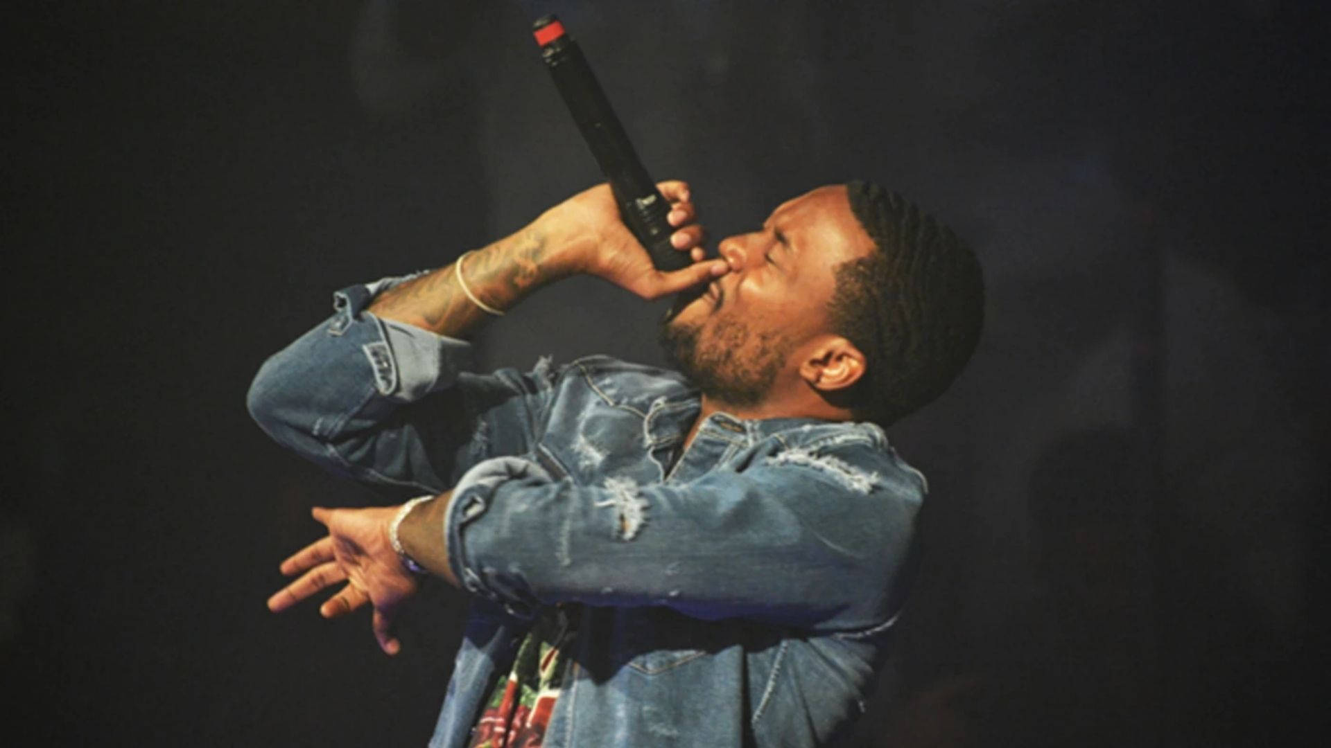 Meek Mill 演唱中壁紙 壁紙 — 一位男性表演者在舞台燈光下熱情地對著麥克風唱歌，身穿破舊的牛仔外套。
