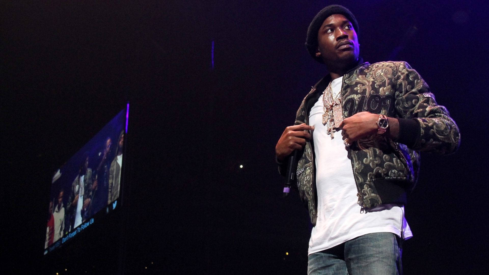 Meek Mill 演唱會現場壁紙 壁紙 — 一位身穿圖案外套和冷帽的饒舌歌手，在紫色燈光下的黑暗舞台上手持麥克風表演。