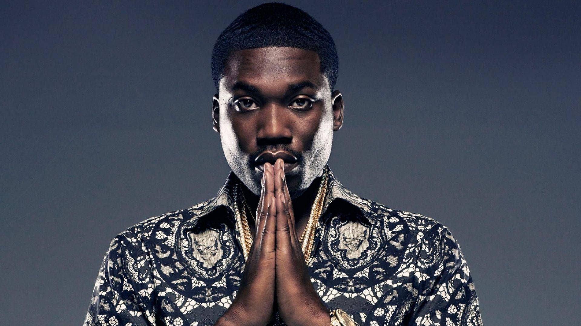 Meek Mill 照片拍攝壁紙 壁紙 — 一名身穿圖案襯衫的男子，雙手合十放在嘴前，背景為深灰色。