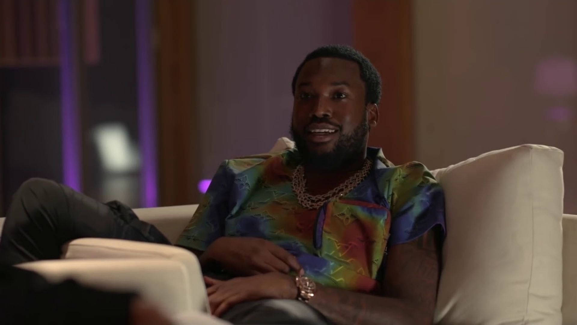Meek Mill 穿彩色襯衫 壁紙 — 一名身穿鮮豔彩虹襯衫的男子，在昏暗的房間裡，紫色環境燈光下，舒適地躺在白色沙發上。