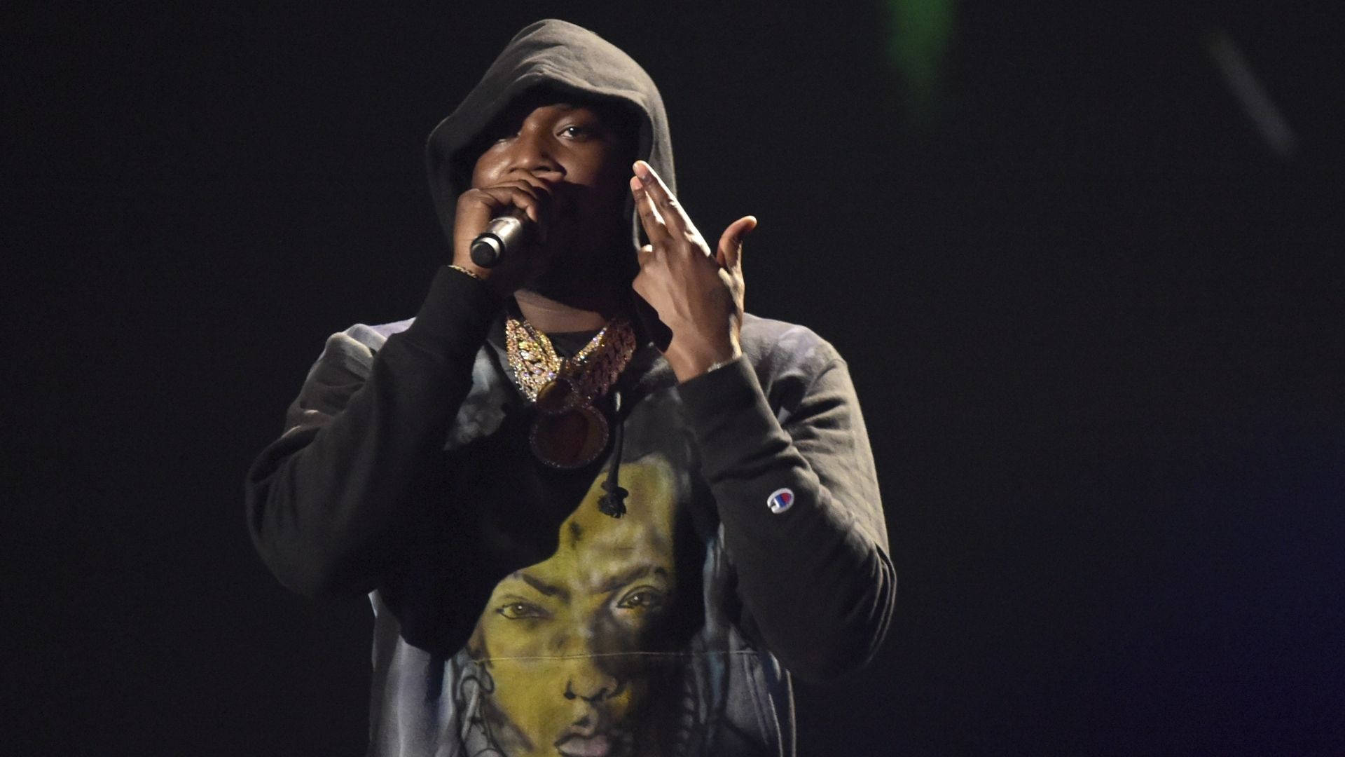 Meek Mill 穿連帽衫 壁紙 — 一位身穿帶有臉部圖案的深色連帽衫的說唱歌手在戲劇性燈光下拿著麥克風在舞台上表演。