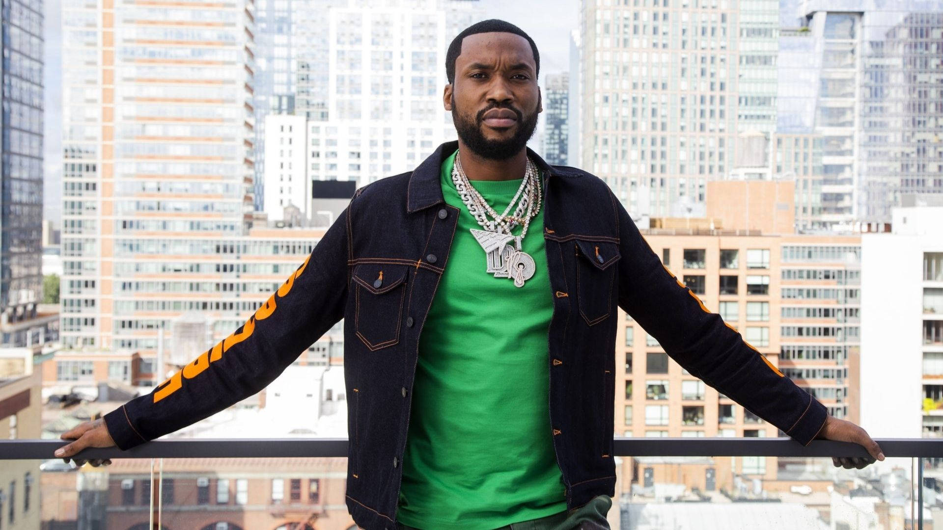 Meek Mill 站在天台 壁紙 — 一名身穿綠色襯衫和黑色牛仔褸（橙色袖）的男子自信地站在城市陽台上，俯瞰摩天大樓。