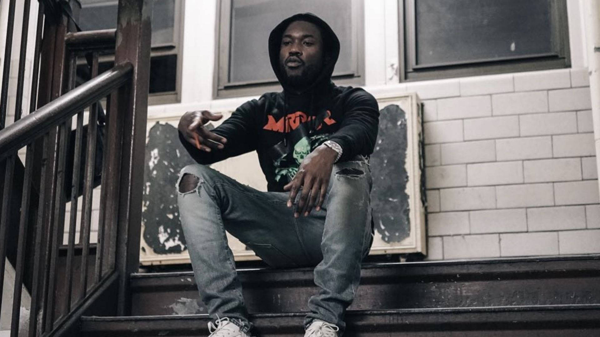 Meek Mill 站階梯 壁紙 — 一個穿著黑色連帽衫和破洞牛仔褲的男人隨意地坐在城市樓梯上，身後有塗鴉和磚牆。