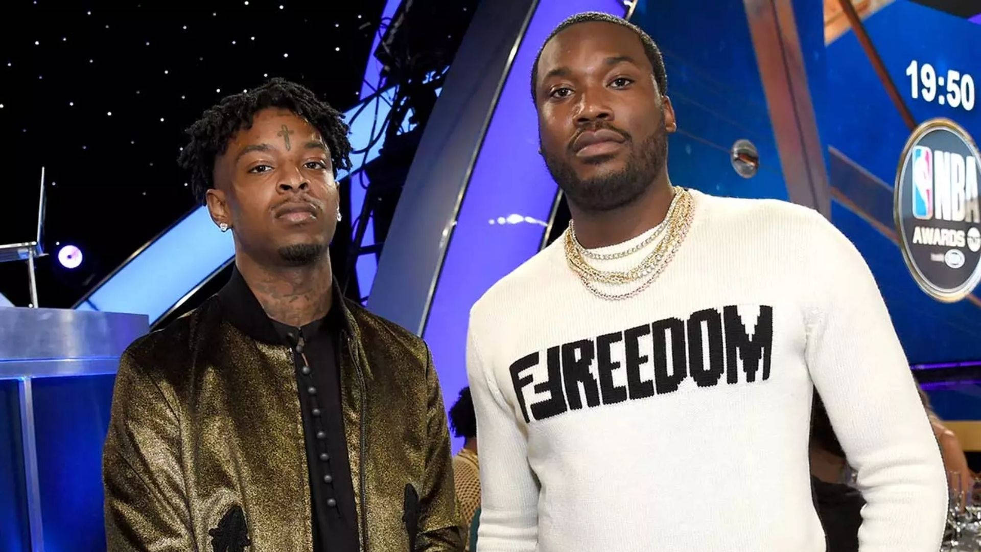 Meek Mill 與 21 Savage 壁紙 — 兩個男人站在藍色燈光的NBA頒獎舞台上，一個穿著金色外套，另一個穿著白色「FREEDOM」毛衣。