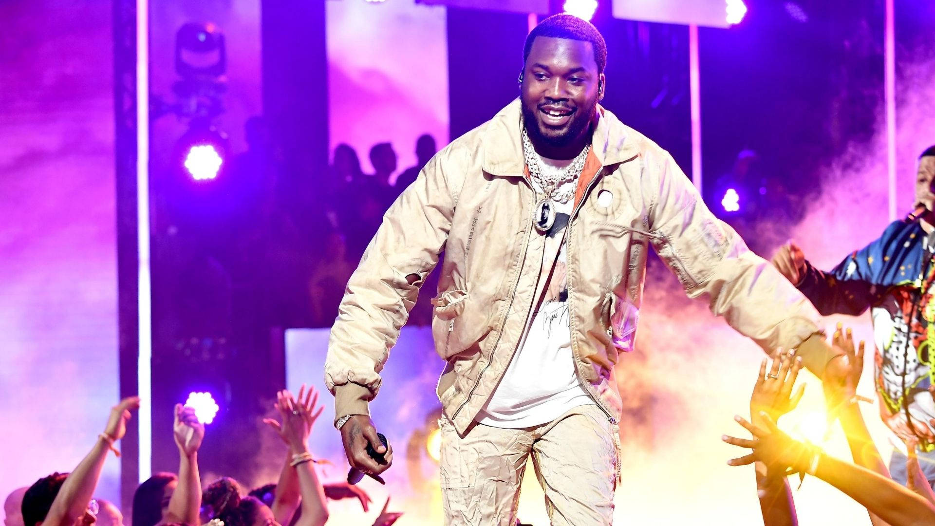 Meek Mill 與粉絲互動 壁紙 — 一位微笑的饒舌歌手穿著米色外套，在紫色燈光的舞台上充滿活力地表演，粉絲們舉起發光的螢光棒。