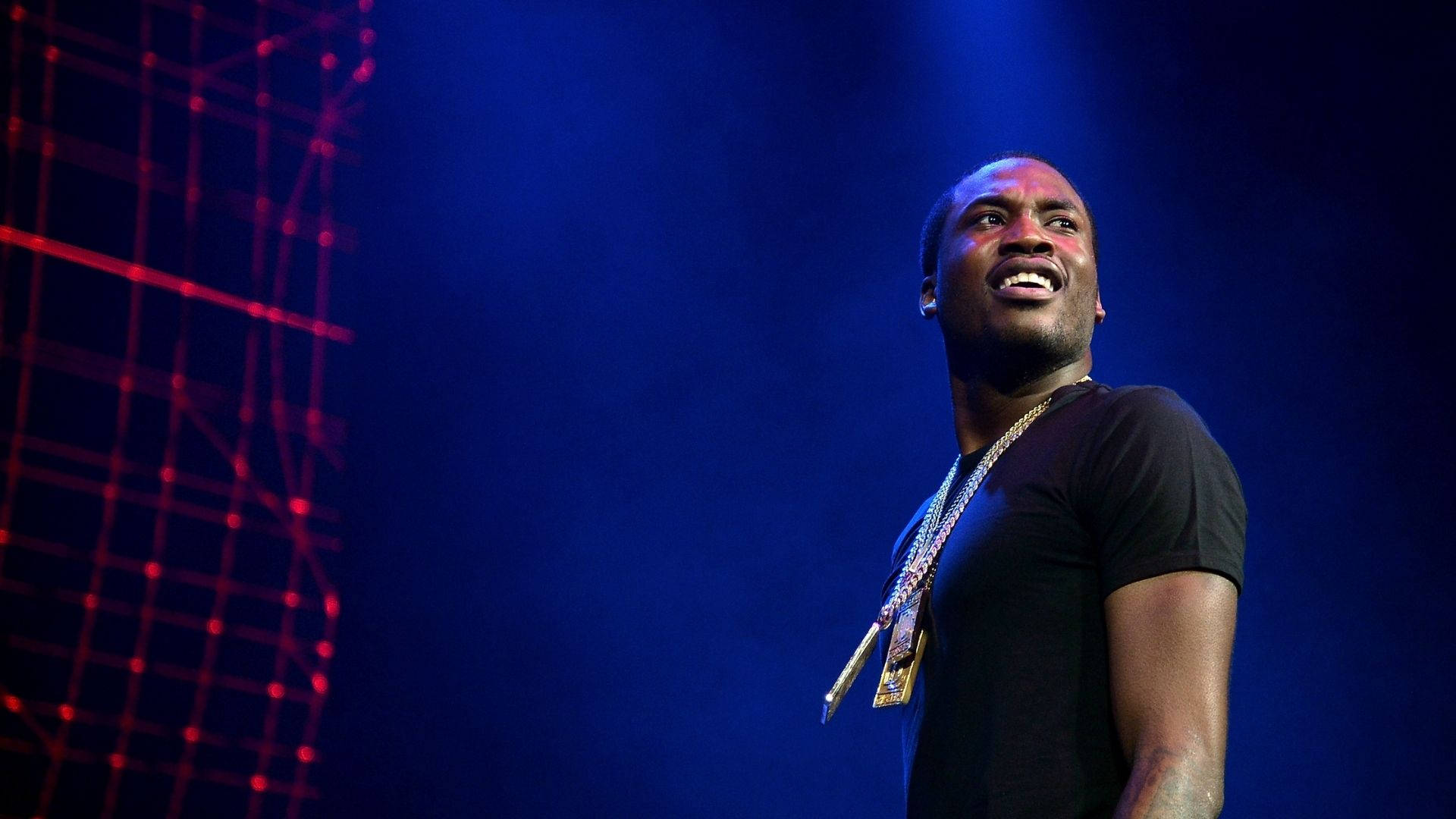 Meek Mill 舞台互動壁紙 壁紙 — 一位微笑的表演者身穿黑色襯衫和金色鏈條，站在被藍色燈光籠罩的舞台上，身後有紅色網格圖案。
