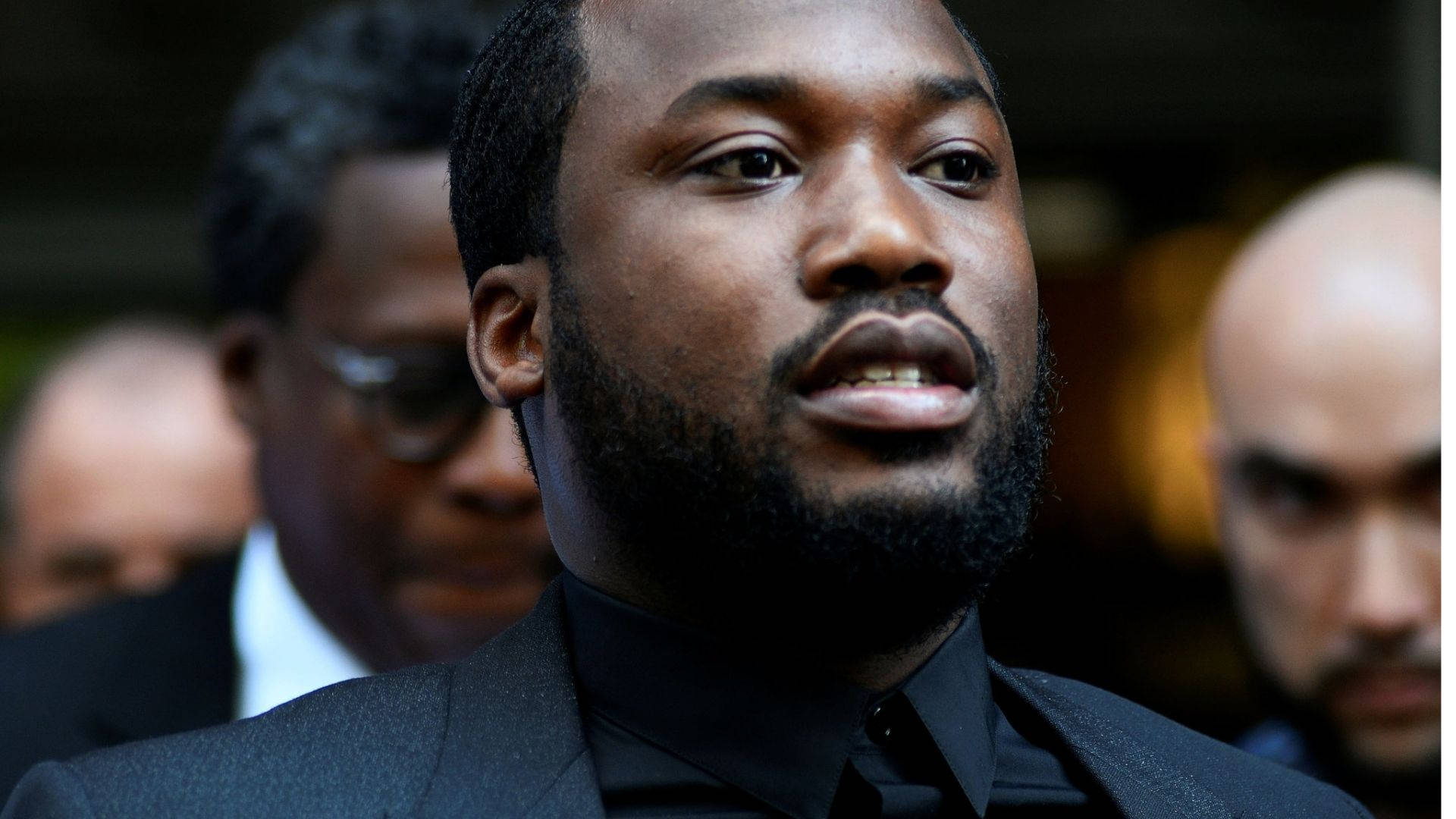 Meek Mill 著黑色西裝 壁紙 — 一位留鬍鬚、身穿黑色西裝和襯衫的男士的特寫肖像，表情嚴肅地向上看。