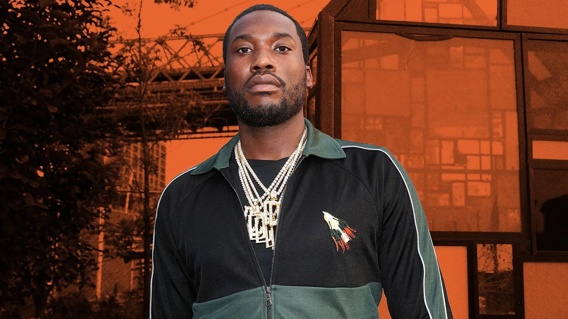 Meek Mill 街頭風采 壁紙 — 一名身穿黑綠色運動外套、佩戴多層金鏈的男子，自信地站在橙色调的城市建築背景下。