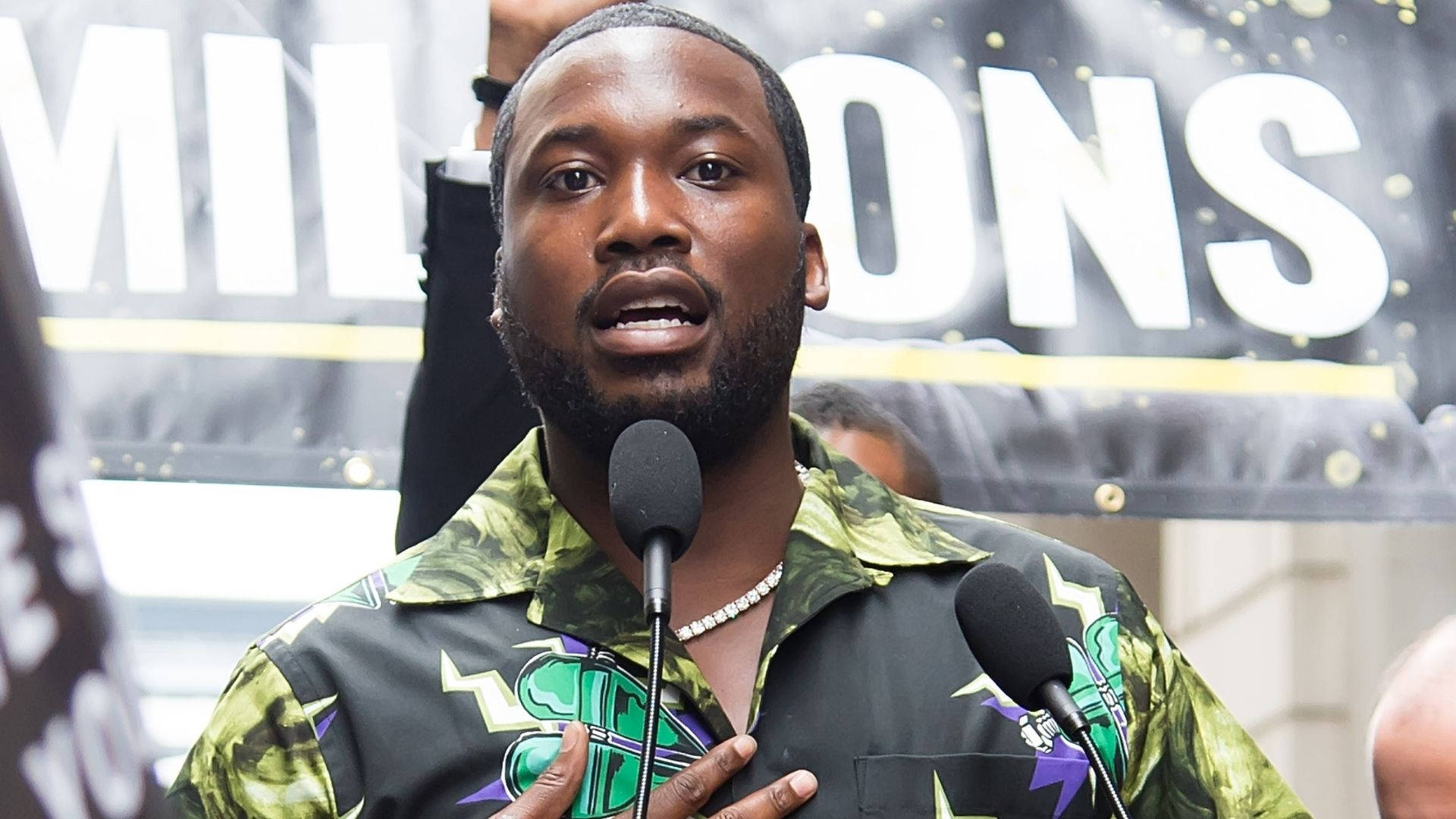 Meek Mill 講話中 壁紙 — 一名身穿鮮豔綠色圖案襯衫的男子在戶外活動中，身後有橫幅，認真地對著麥克風說話。