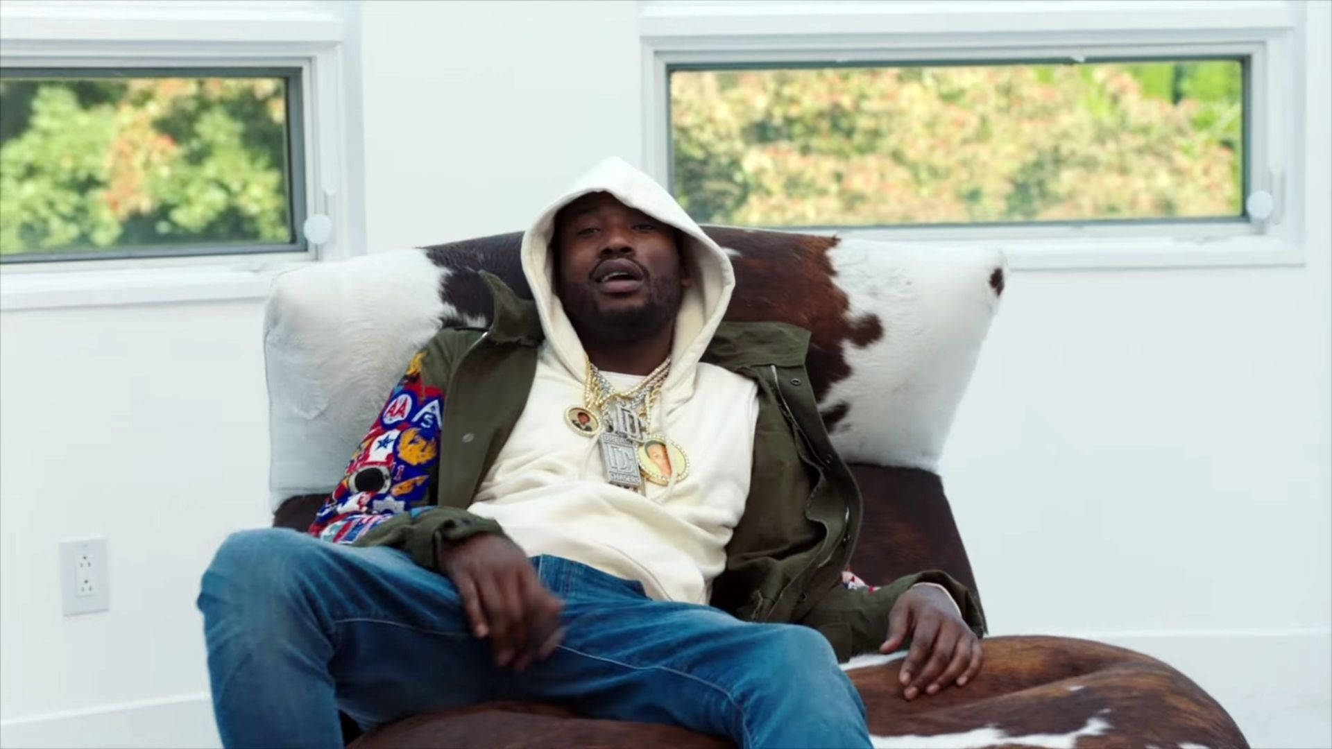 Meek Mill 躺沙發 壁紙 — 一名身穿白色連帽衫和綠色外套的男子，在一間明亮的白色房間裡，躺在牛毛椅上，窗外可見綠意。