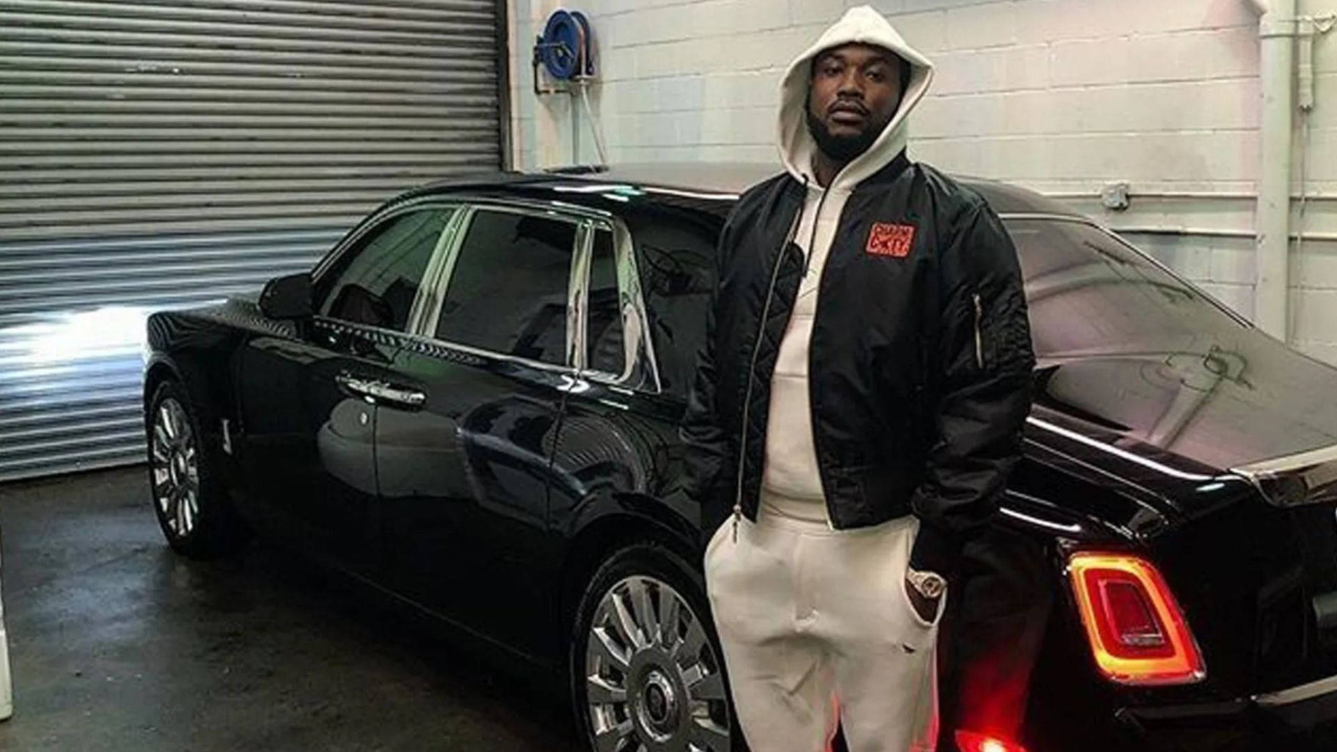Meek Mill 車庫 壁紙 — 一名身穿白色連帽衫和黑色外套的男子自信地站在一輛光澤黑色勞斯萊斯旁邊，身後是帶有金屬門的混凝土車庫。