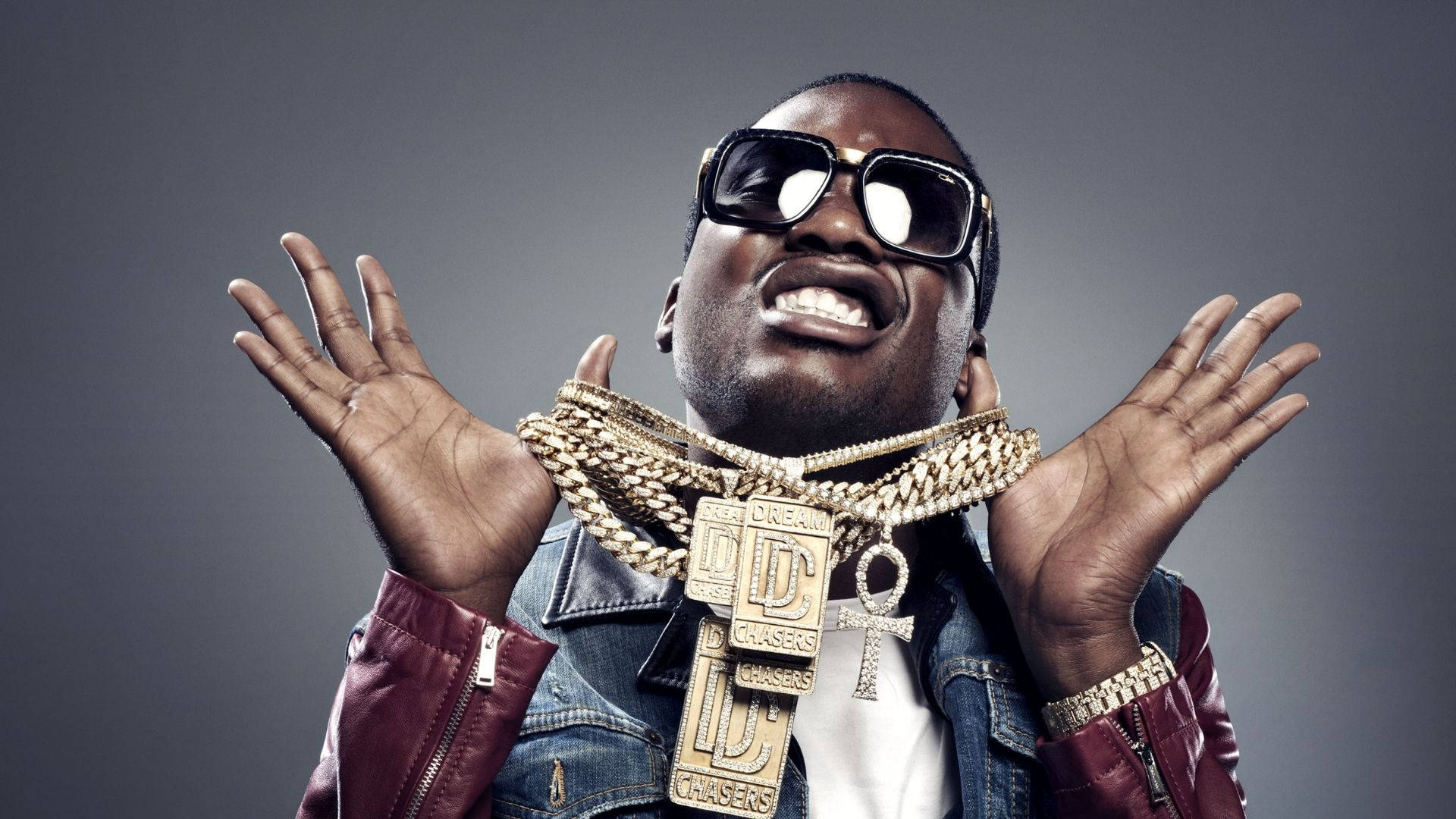 Meek Mill 金飾炫目壁紙 壁紙 — 一名戴著太陽眼鏡和金色鏈條的男子在灰色攝影棚背景下舉起雙手開懷大笑。