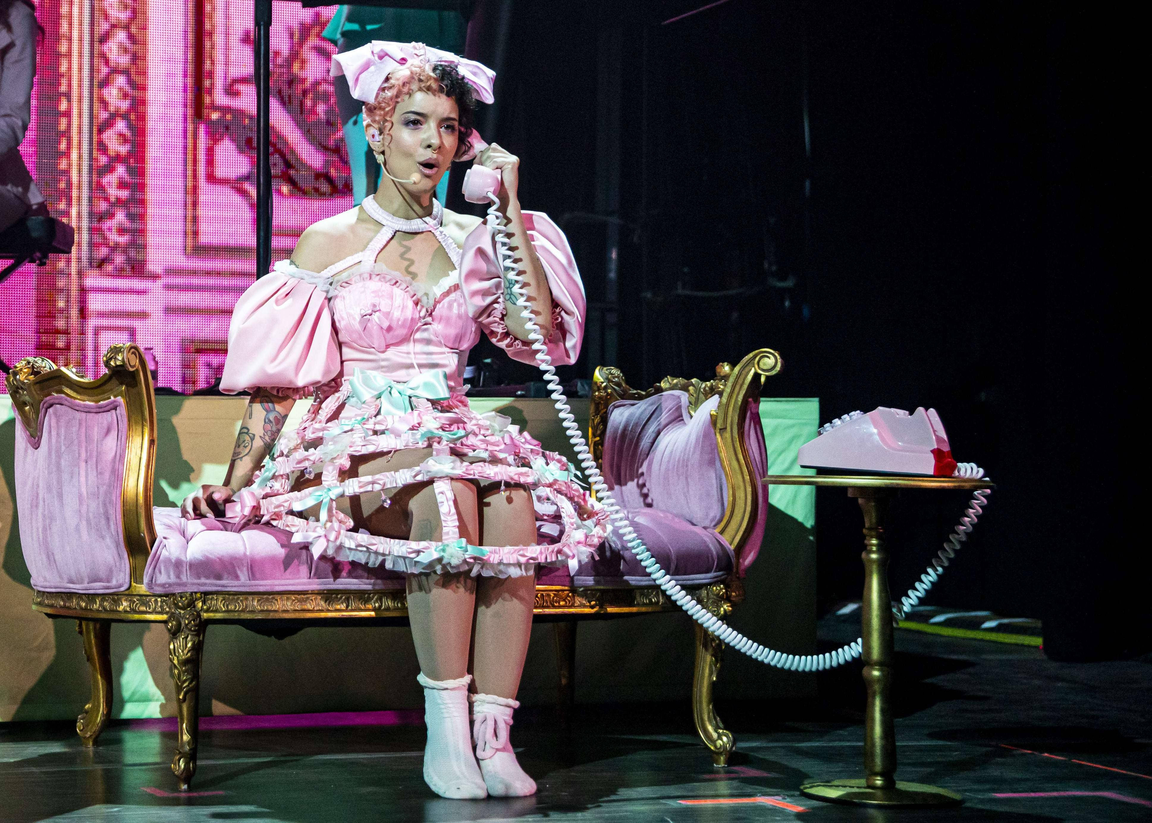 Melanie Martinez 粉紅電話 壁紙 — 一位穿著粉紅色荷葉邊女傭服的表演者坐在鍍金長椅上，在粉紅色燈光下拿著復古電話。