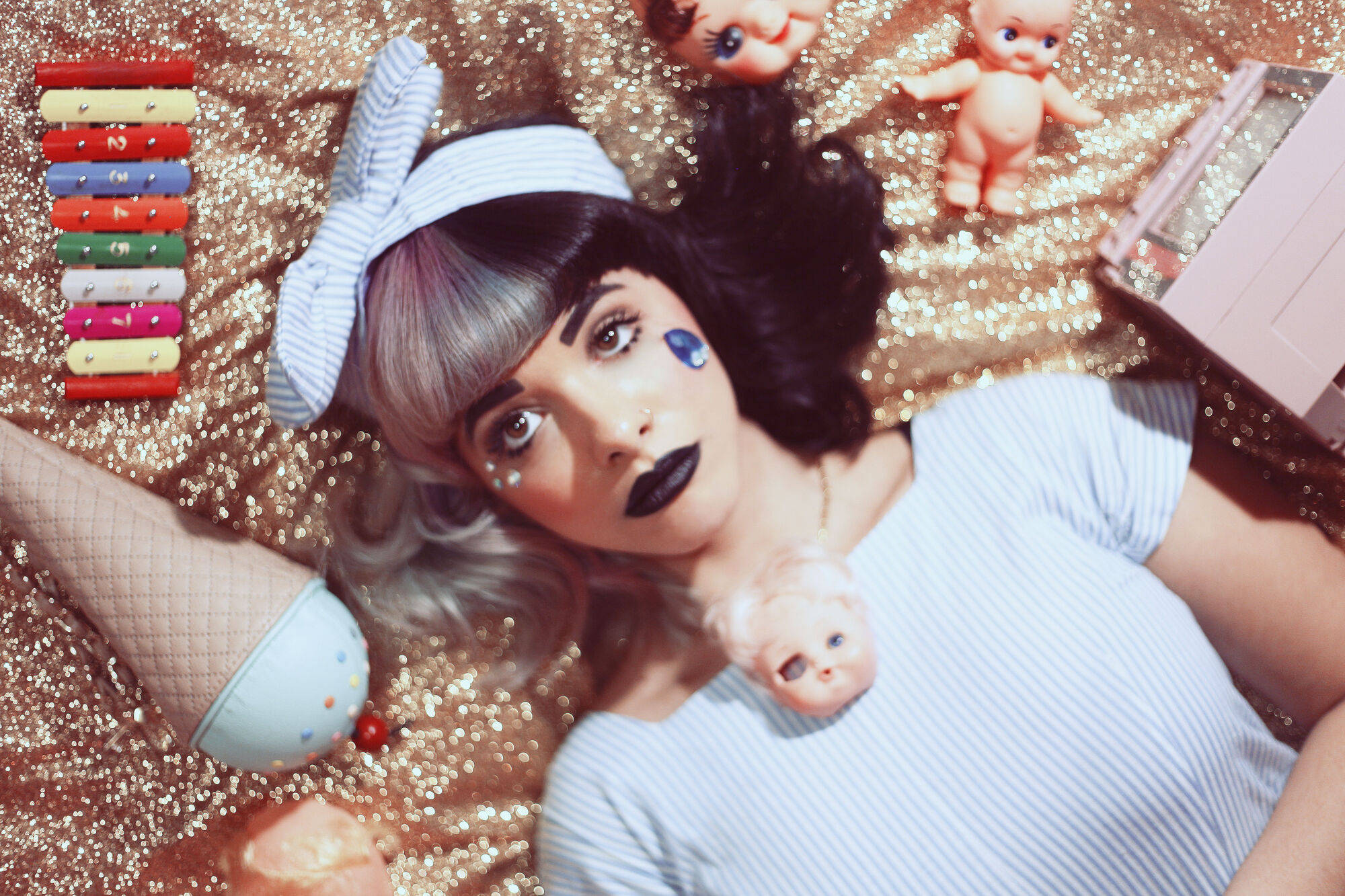 Melanie Martinez 閃耀 壁紙 — 一位塗著深色唇膏、戴著條紋蝴蝶結的女性躺在閃閃發光的金色布料上，周圍環繞著洋娃娃和一個色彩繽紛的木琴。