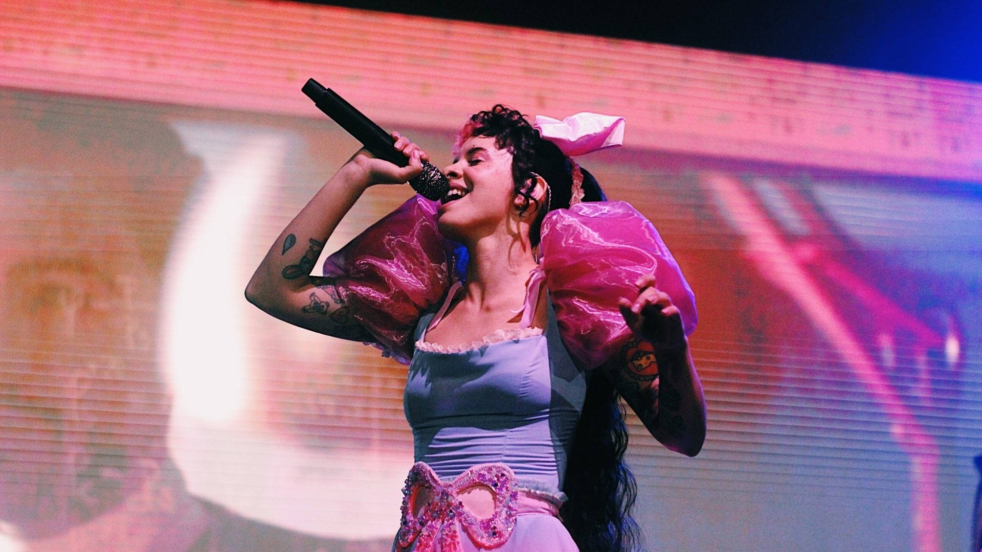 Melanie Martinez 粉紅蓬袖 壁紙 — 一位帶有紋身和粉紅色蝴蝶結的歌手在充滿活力的粉紅色和藍色燈光下充滿活力地在舞台上表演。
