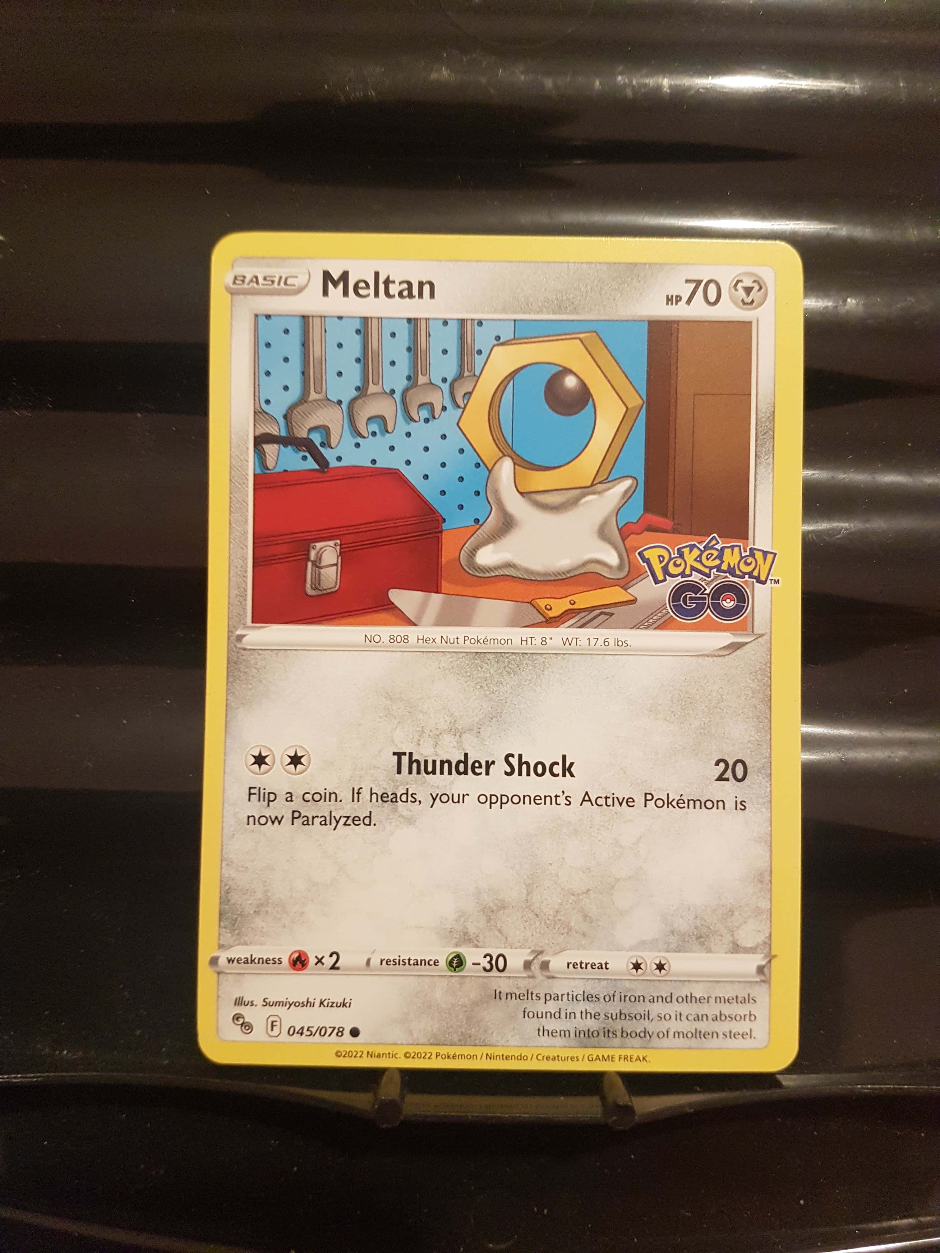 Meltan 寶可夢卡牌 壁紙 — 一張寶可夢卡牌，描繪美錄坦在工作室中，周圍有工具和一個紅色工具箱，背景是藍色圓點牆。