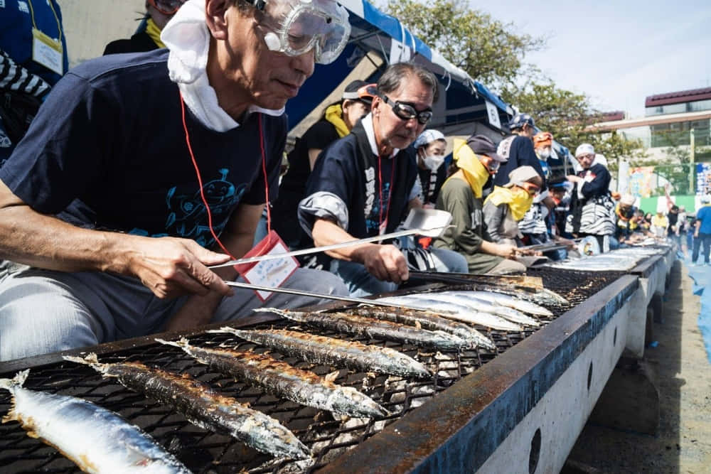 Pacific Saury 桌布 壁紙 — 男士們在陽光明媚的節日裡，在長長的戶外烤架上烤長魚，有觀眾觀看。