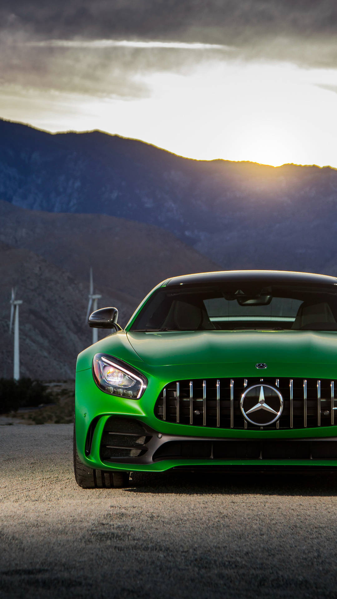 綠色 Mercedes-AMG 跑車 iPhone 壁紙 — 一輛充滿活力的綠色 Mercedes-AMG GT 跑車停在碎石路上，背景是風力發電機和山脈，天空呈現戲劇性的日落景色。