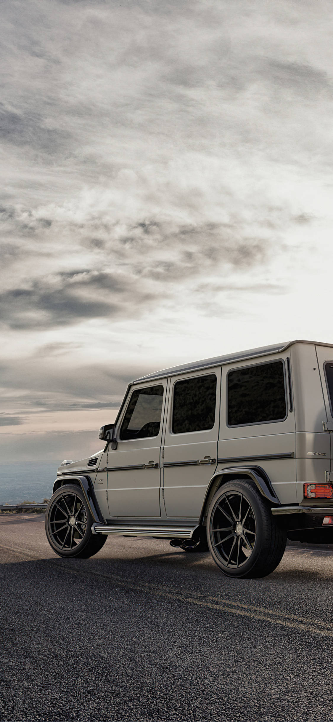 Mercedes Benz 白色 G Wagon iPhone 壁紙 — 一架銀色 Mercedes G-Wagen 停泊在海岸公路上，天空烏雲密布，氣氛戲劇化。