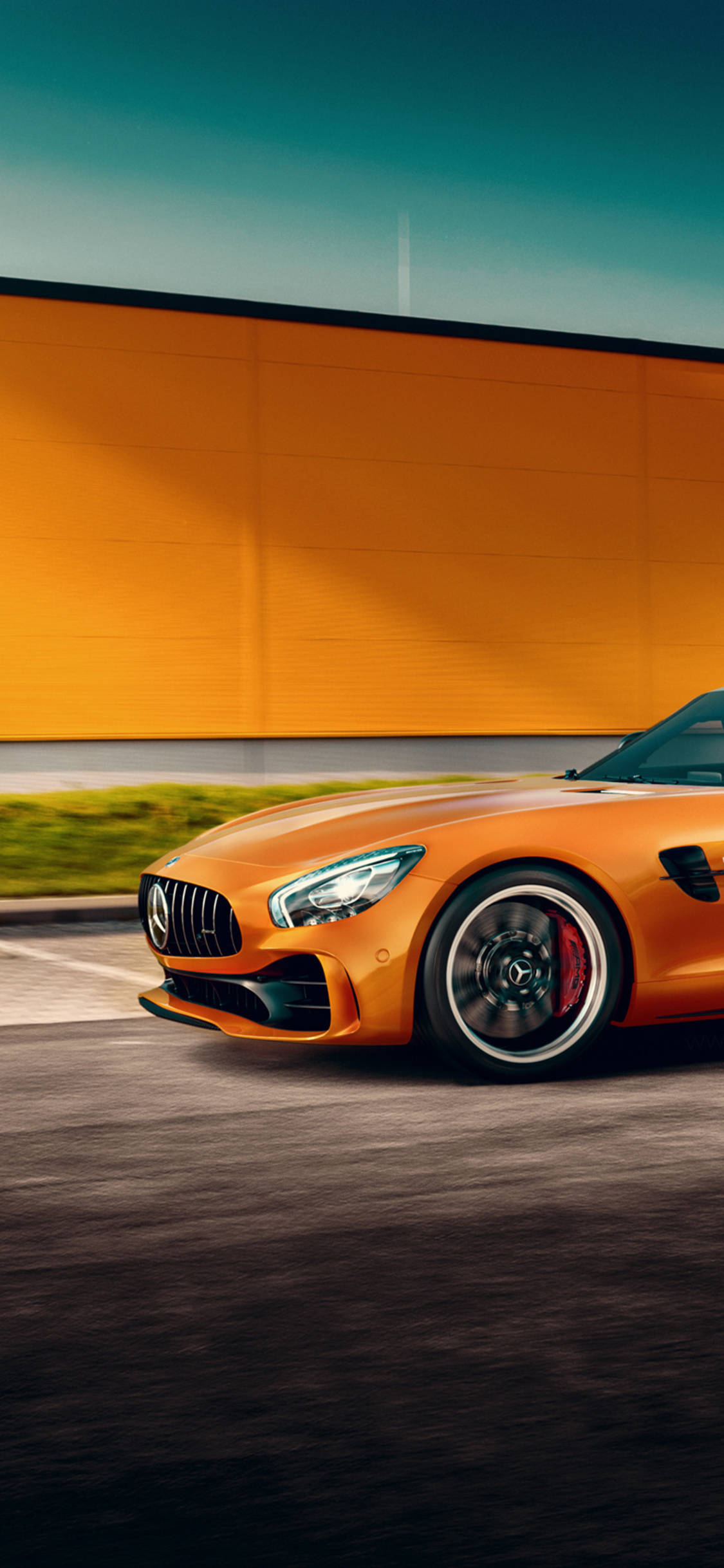橙色Mercedes iPhone X 壁紙 — 一輛充滿活力的橙色 Mercedes-AMG GT 跑車在晴朗的藍天下飛馳而過一道大膽的橙色牆壁。