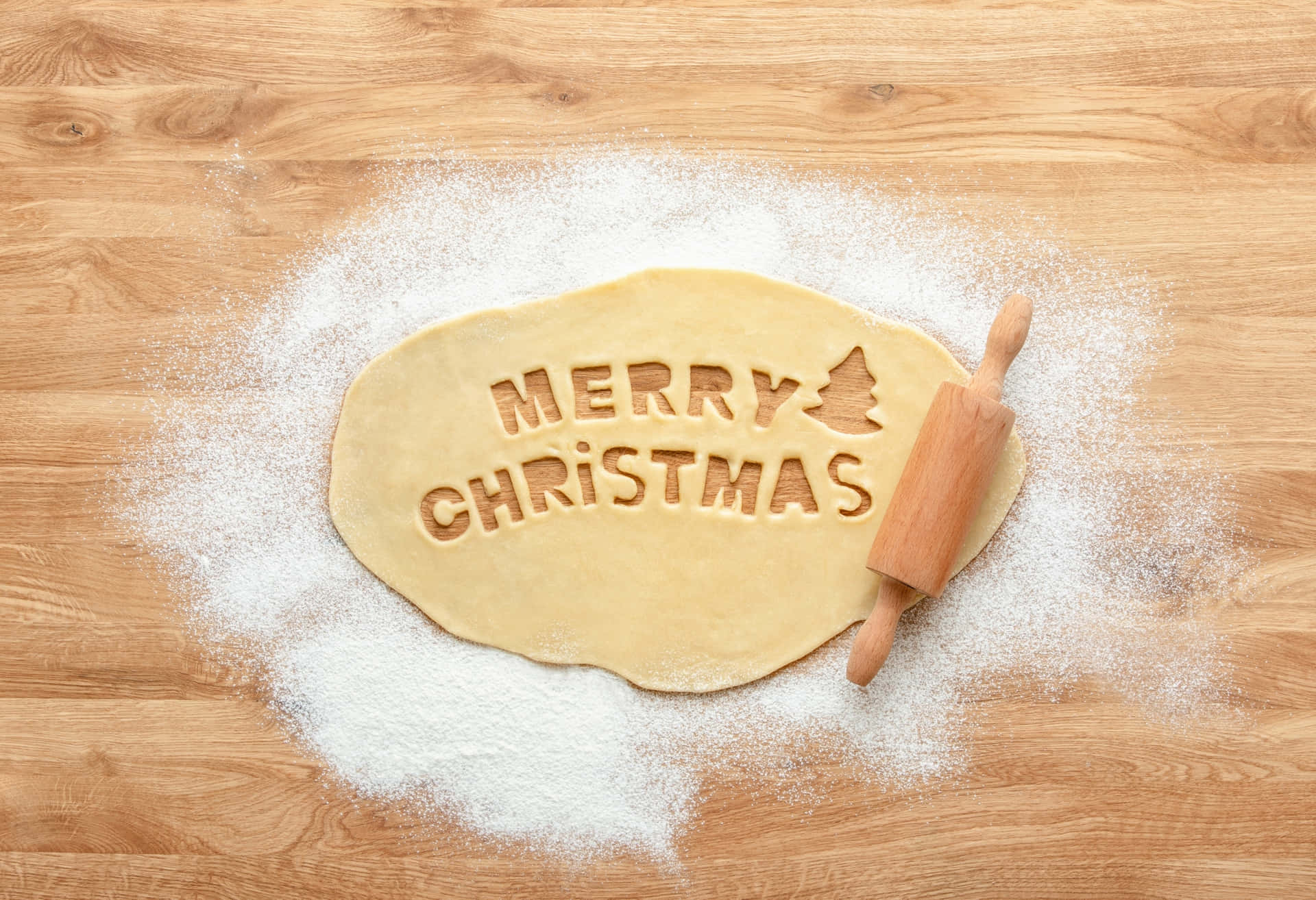 麵糰上印有「Merry Christmas」和樹的圖案，在木桌上用麵粉呈現，旁邊有擀麵杖。