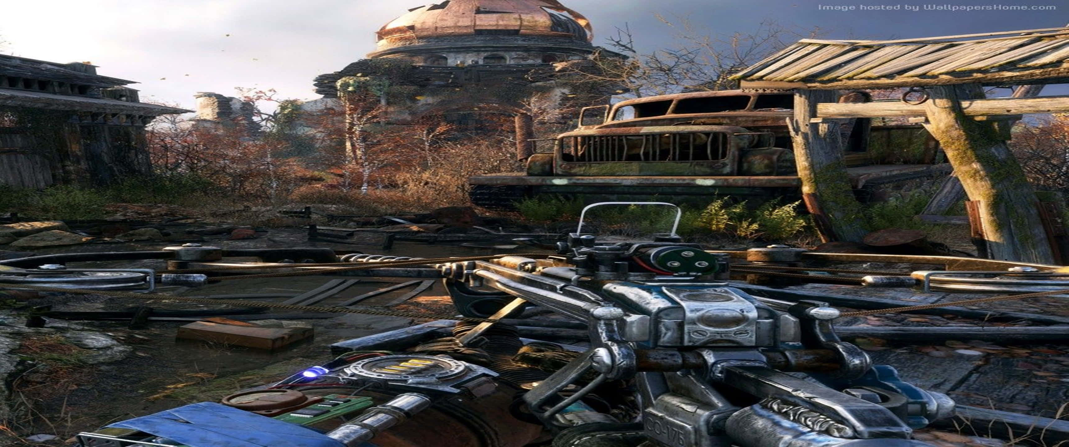 《Metro Exodus》弩箭武器 3440x1440p遊戲背景 壁紙 — 在一個破敗的後末日定居點廢墟中，生鏽的車輛和雜草叢生的建築物在朦朧的天空下，一把未來武器的第一人稱視角。