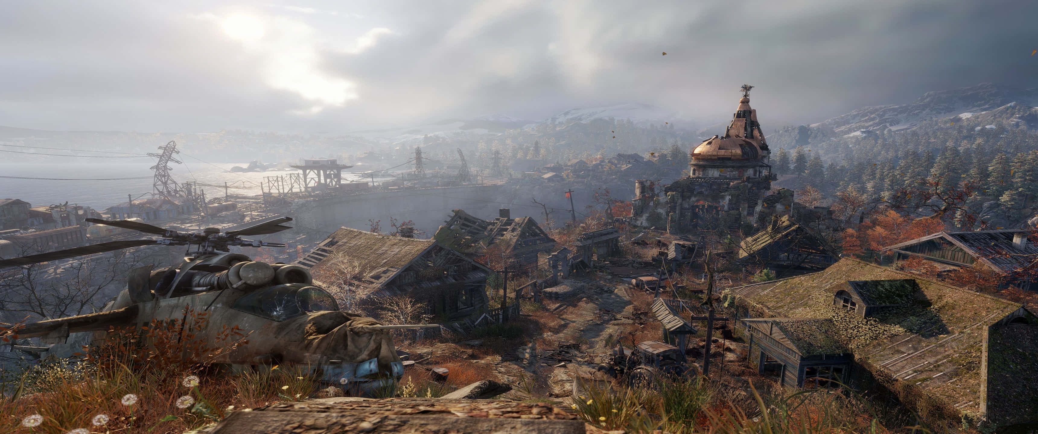 《Metro Exodus》村莊鳥瞰圖 3440x1440p 壁紙 — 一個被毀壞的村莊，前景有一架墜毀的直升機，俯瞰著多雲天空下霧氣瀰漫、積雪的工業廢墟。
