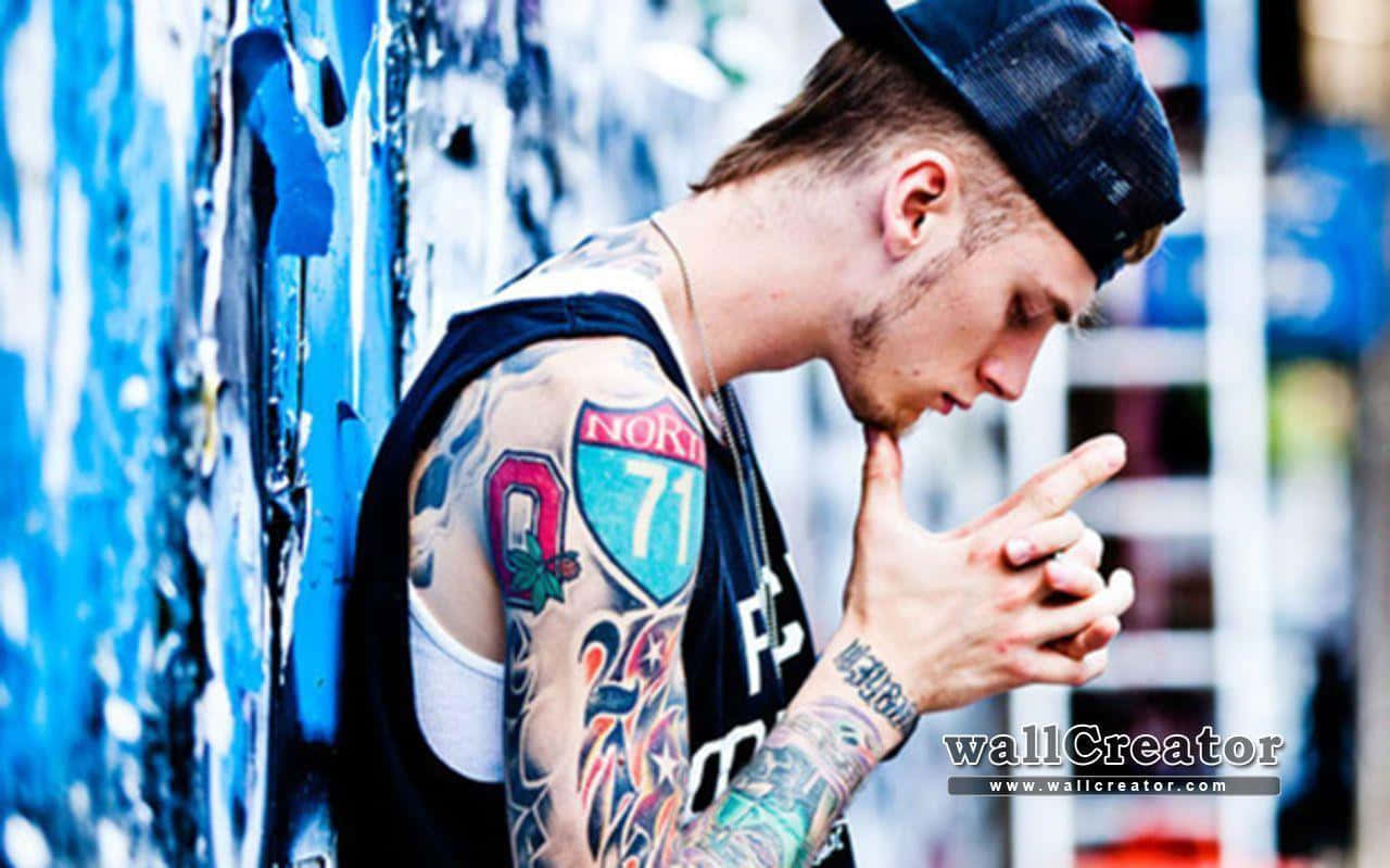 MGK 低頭凝視 壁紙 — 一位紋身青年戴著帽子，靠在一幅佈滿塗鴉的藍色牆上，手指輕觸嘴唇，擺出沉思的姿勢。