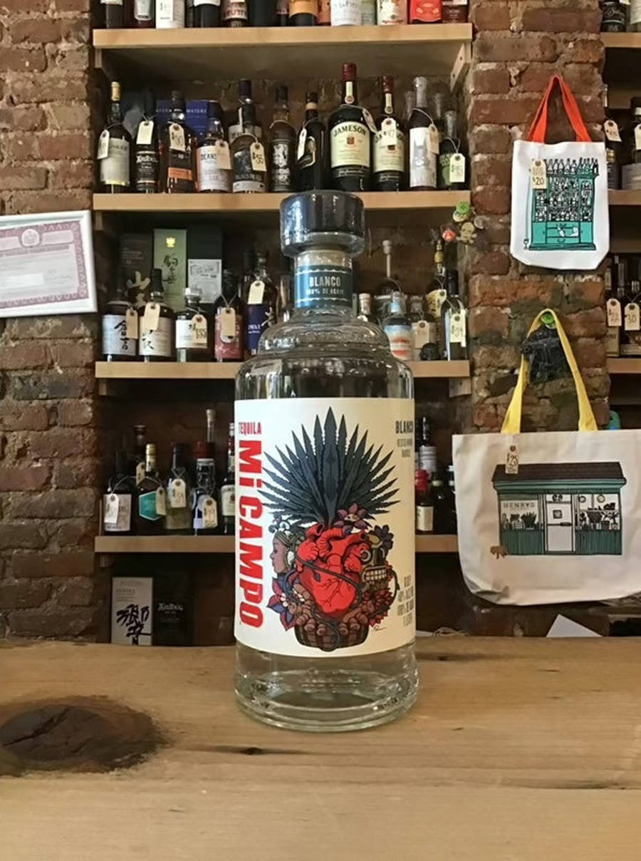 一瓶 Mi Campo 蒂克利亞 750ml 壁紙 — 一瓶透明的Mi Campo龍舌蘭酒，帶有鮮豔的紅色心形和龍舌蘭設計標籤，放在木製吧檯上，背景是磚牆和擺滿酒瓶的架子。